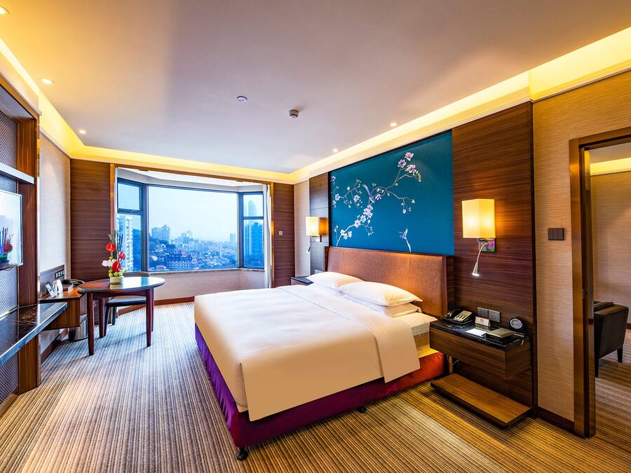Habitación de hotel con una cama grande, un escritorio con silla, una mesita de noche, una lámpara de pared y vistas a la ciudad a través de un ventanal amplio.