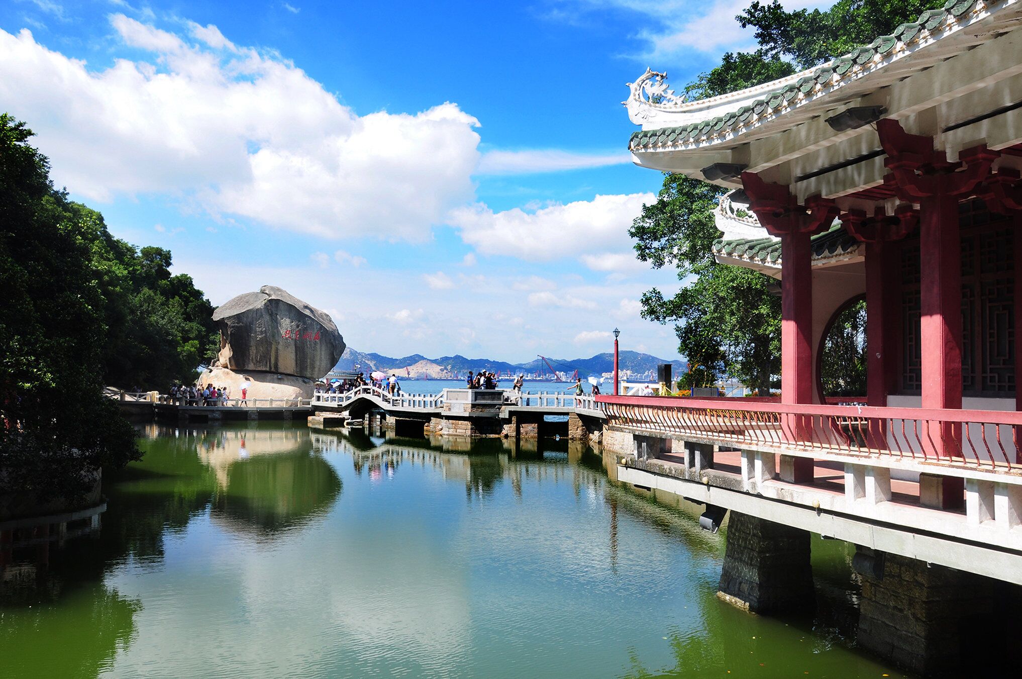 A traditional Chinese pavilion with a reflection in the water, a stone bridge, and a mountainous backdrop.