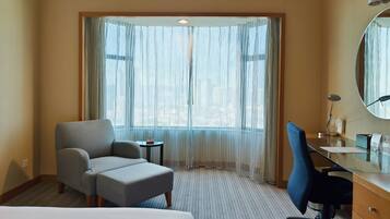 A hotel room with a bed, a desk, a chair, a small table, and a large window with sheer curtains.