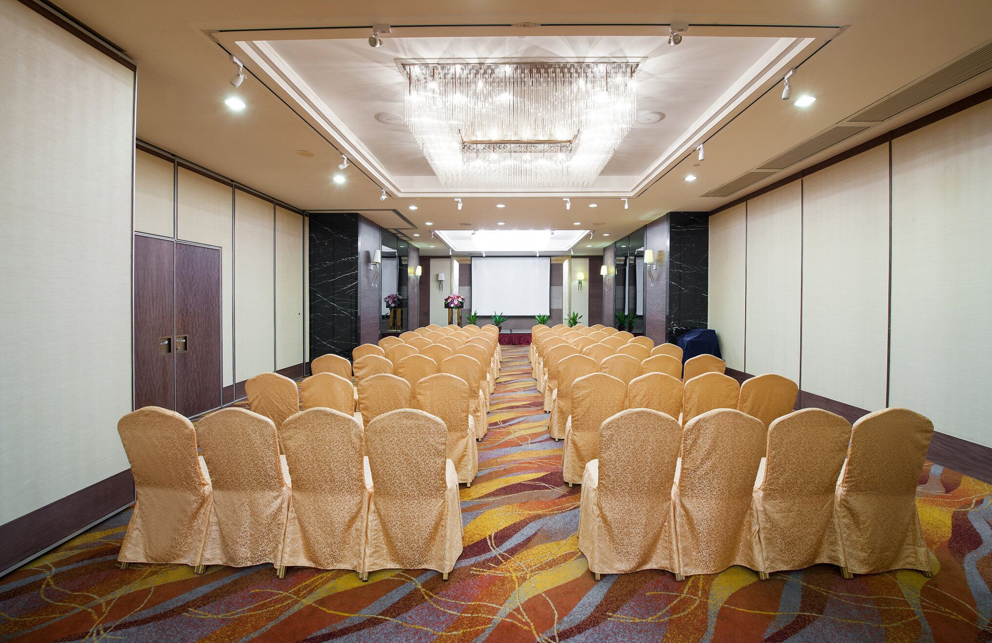 Un salón de conferencias con filas de sillas, una pantalla de proyector y una araña de cristal.