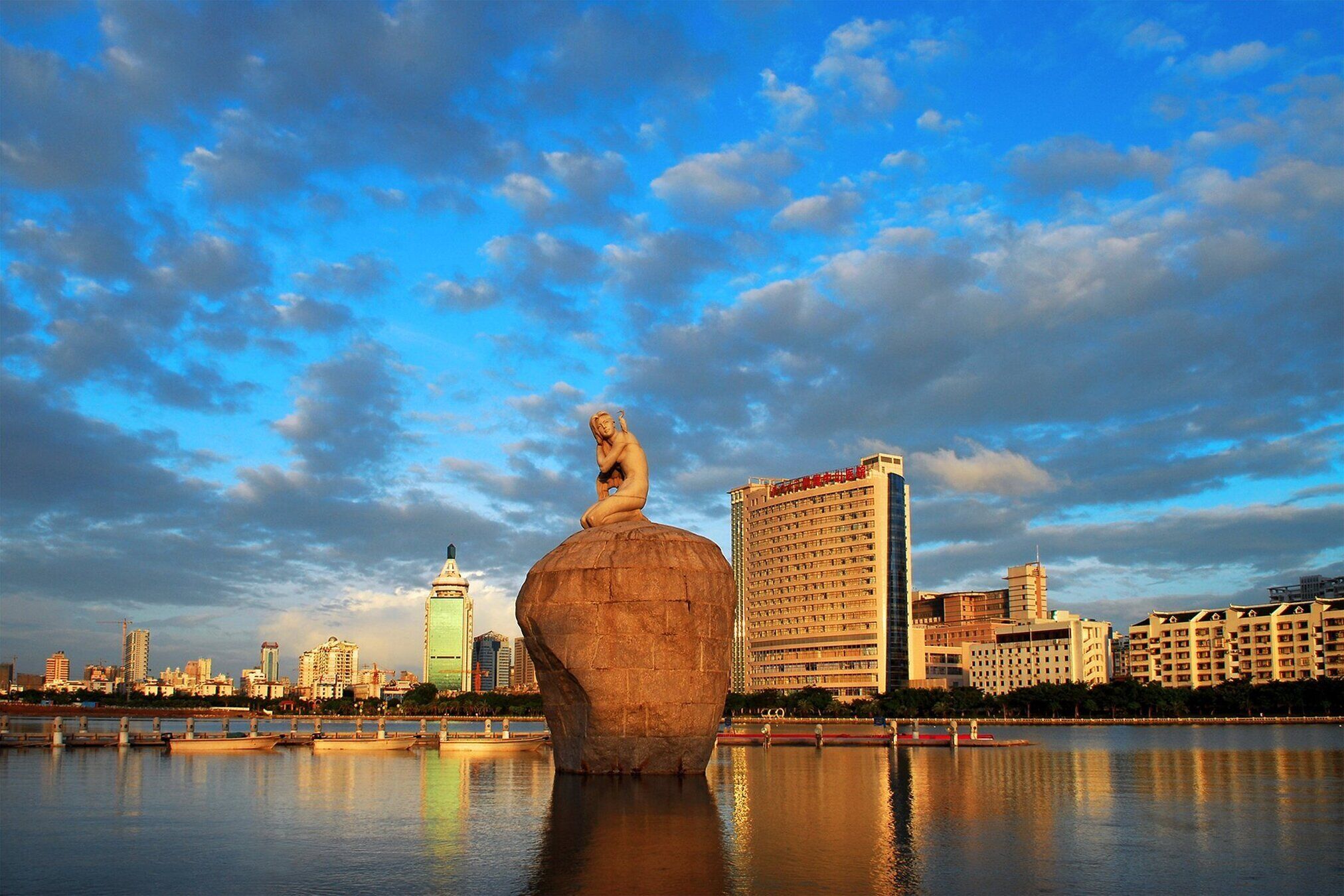 A large stone sculpture in a body of water with a city skyline in the background.