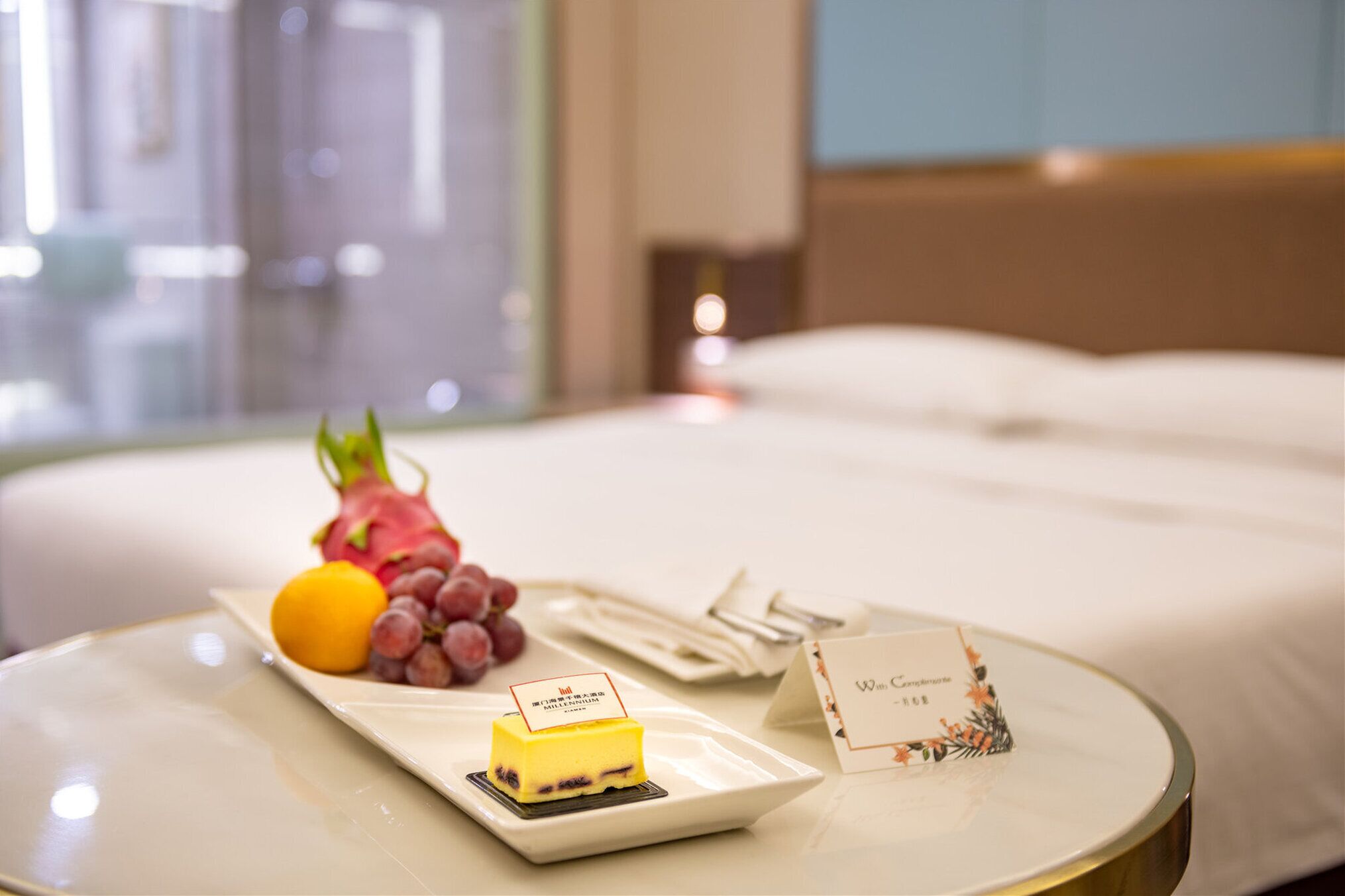 A neatly made bed with a bedside table holding a plate with a dessert, fruit, and a card.