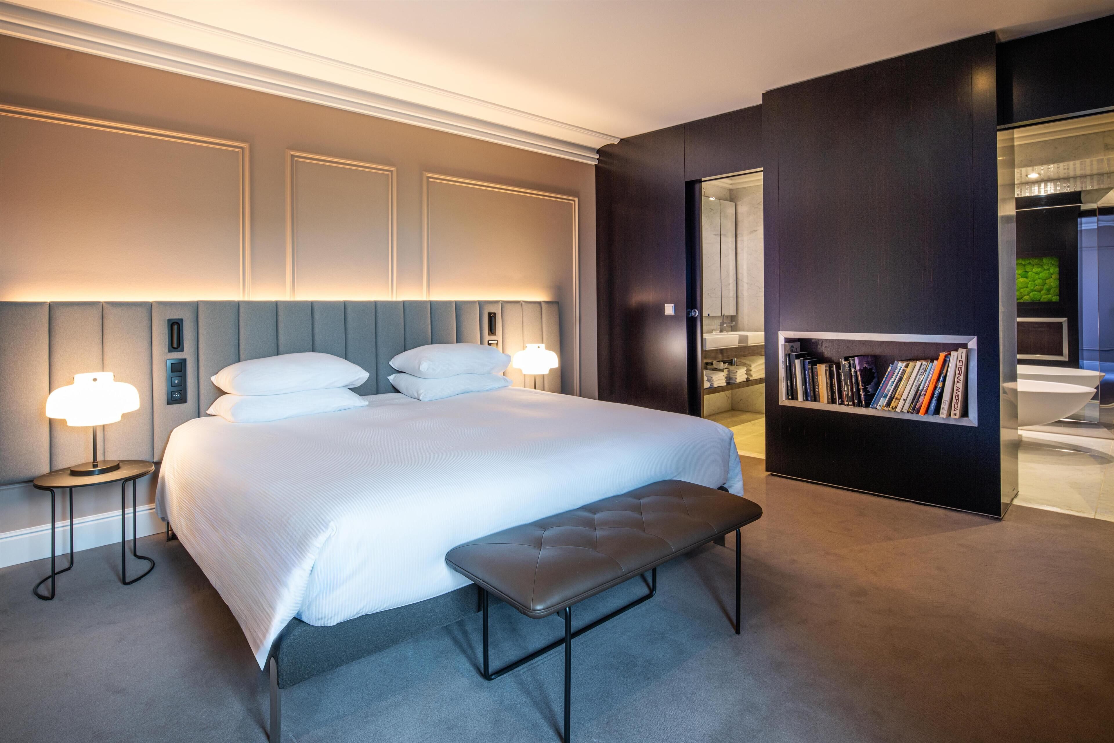 Una habitación de hotel moderna con una cama grande, dos lámparas de noche, un banco y una estantería.