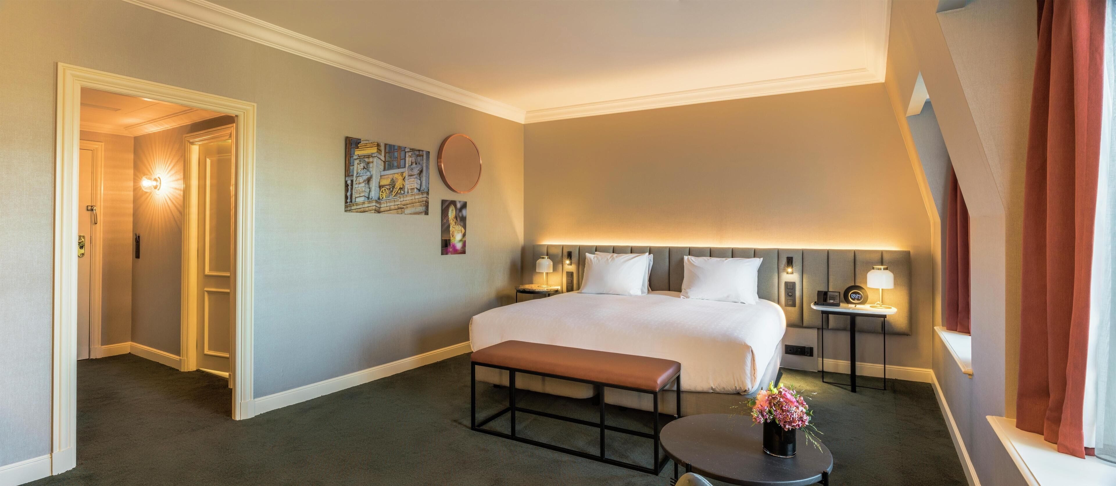 A hotel room with a large bed, bedside tables, a bench, and a small table with a vase of flowers.