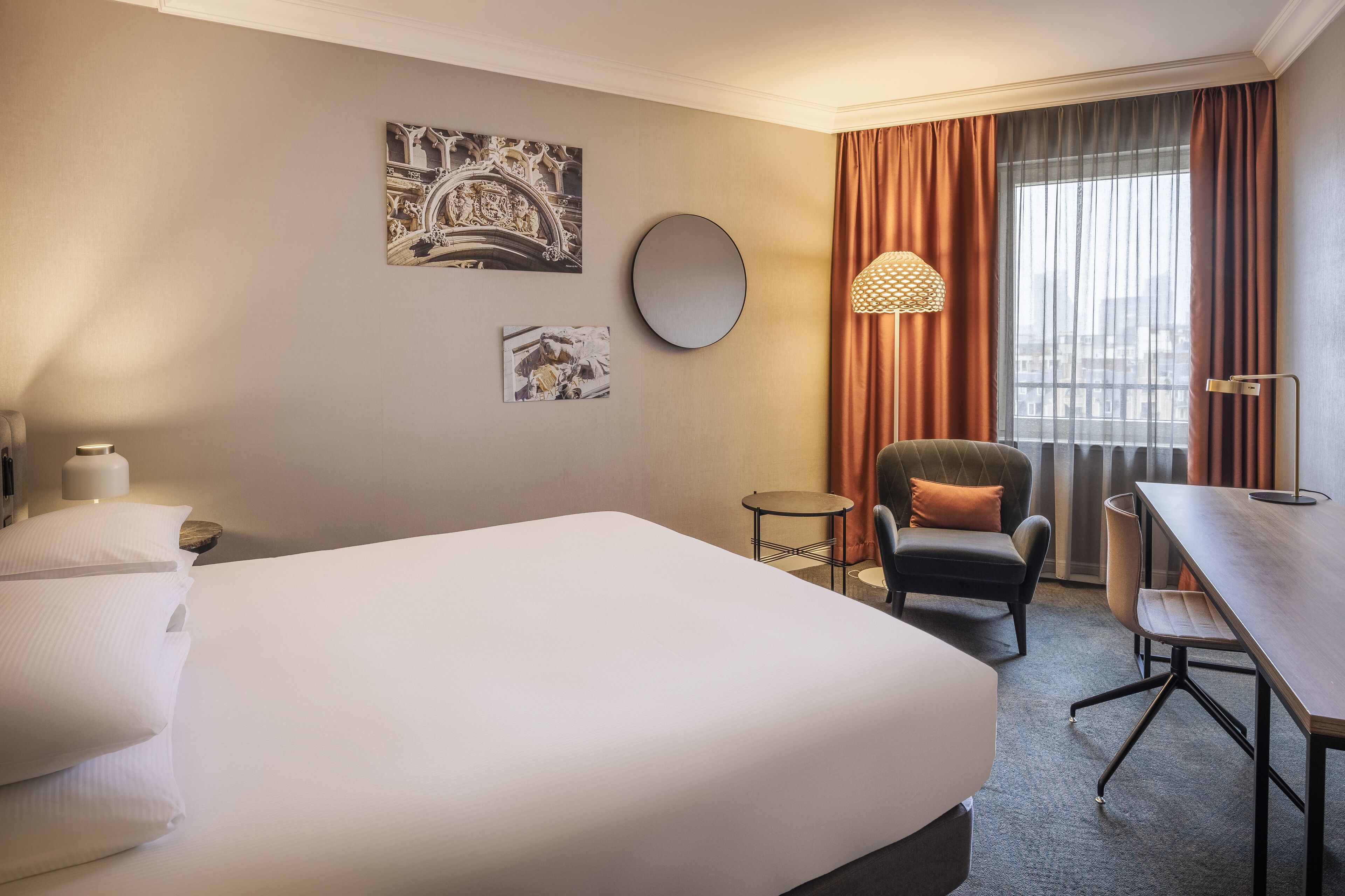 A hotel room with a large bed, a desk, a chair, a round mirror, and two wall-mounted artworks.