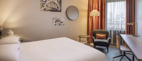 A hotel room with a large bed, a desk, a chair, a round mirror, and two wall-mounted artworks.