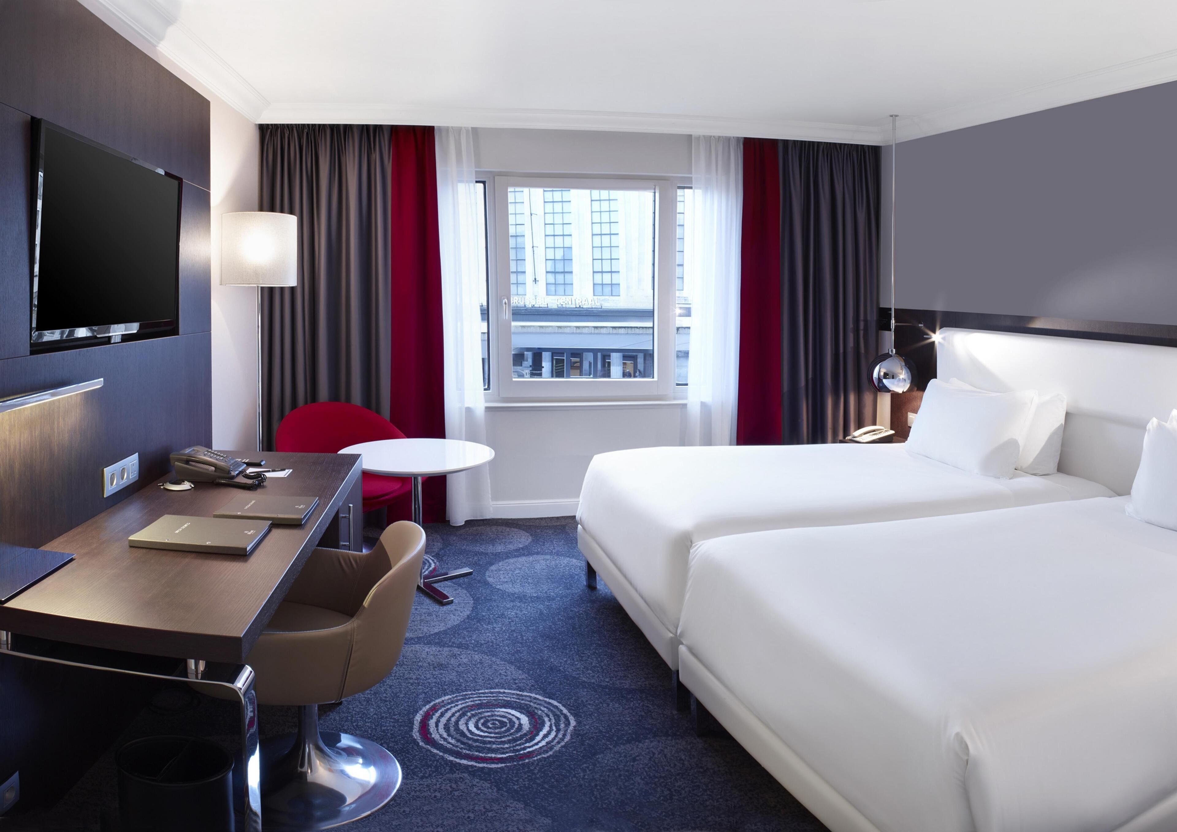 A hotel room with a bed, a desk, a chair, a TV, and a window with curtains.