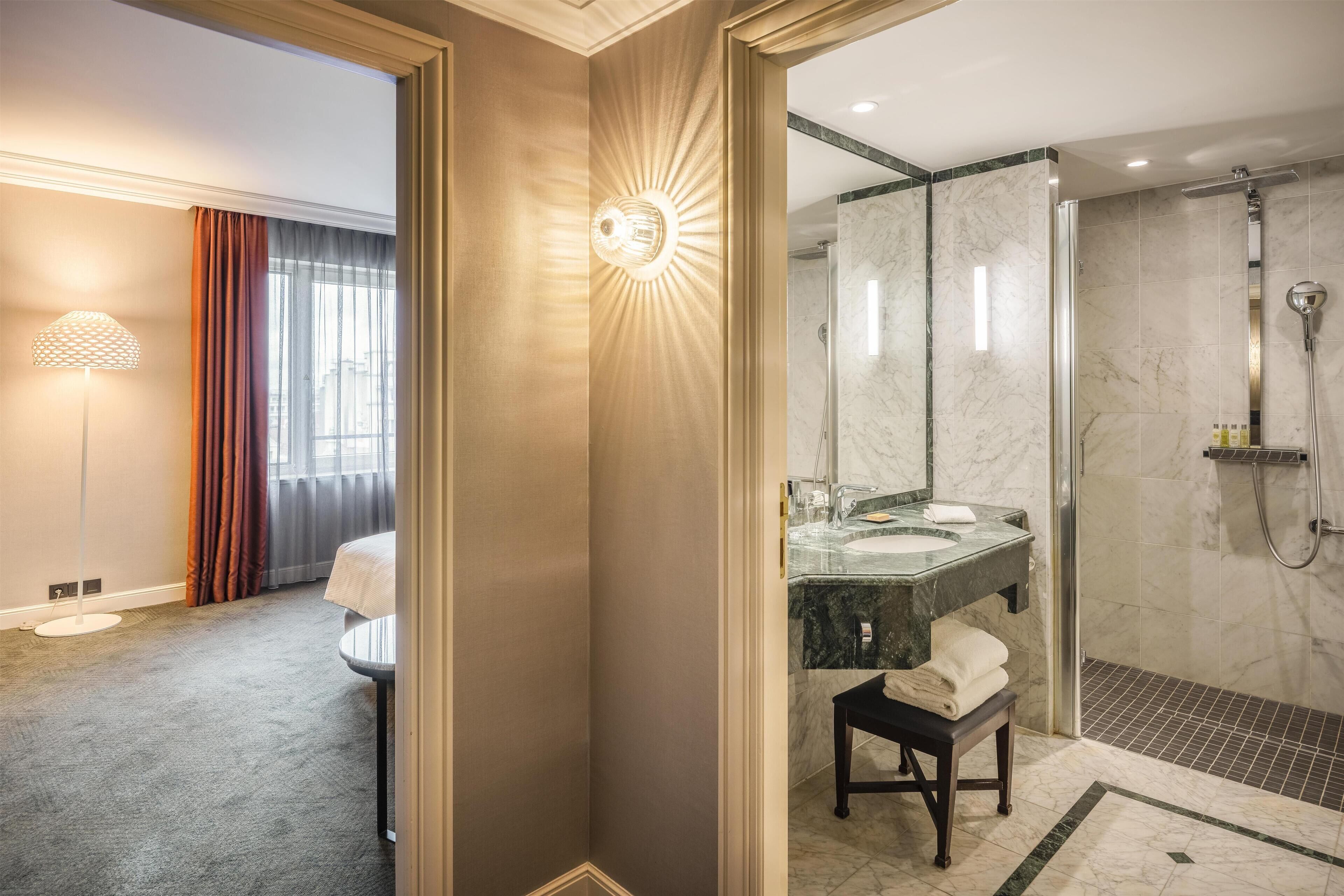 A hotel room with a large mirror, a bedside table, a lamp, and a view of the bathroom.