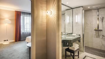 A hotel room with a large mirror, a bedside table, a lamp, and a view of the bathroom.