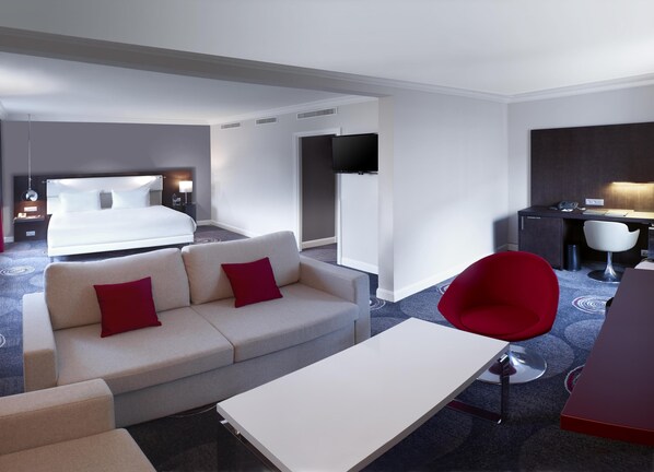 A hotel room with a bed, a sofa with red cushions, a desk with a chair, and a TV mounted on the wall. - Hilton Brussels Grand Place (Brussels)