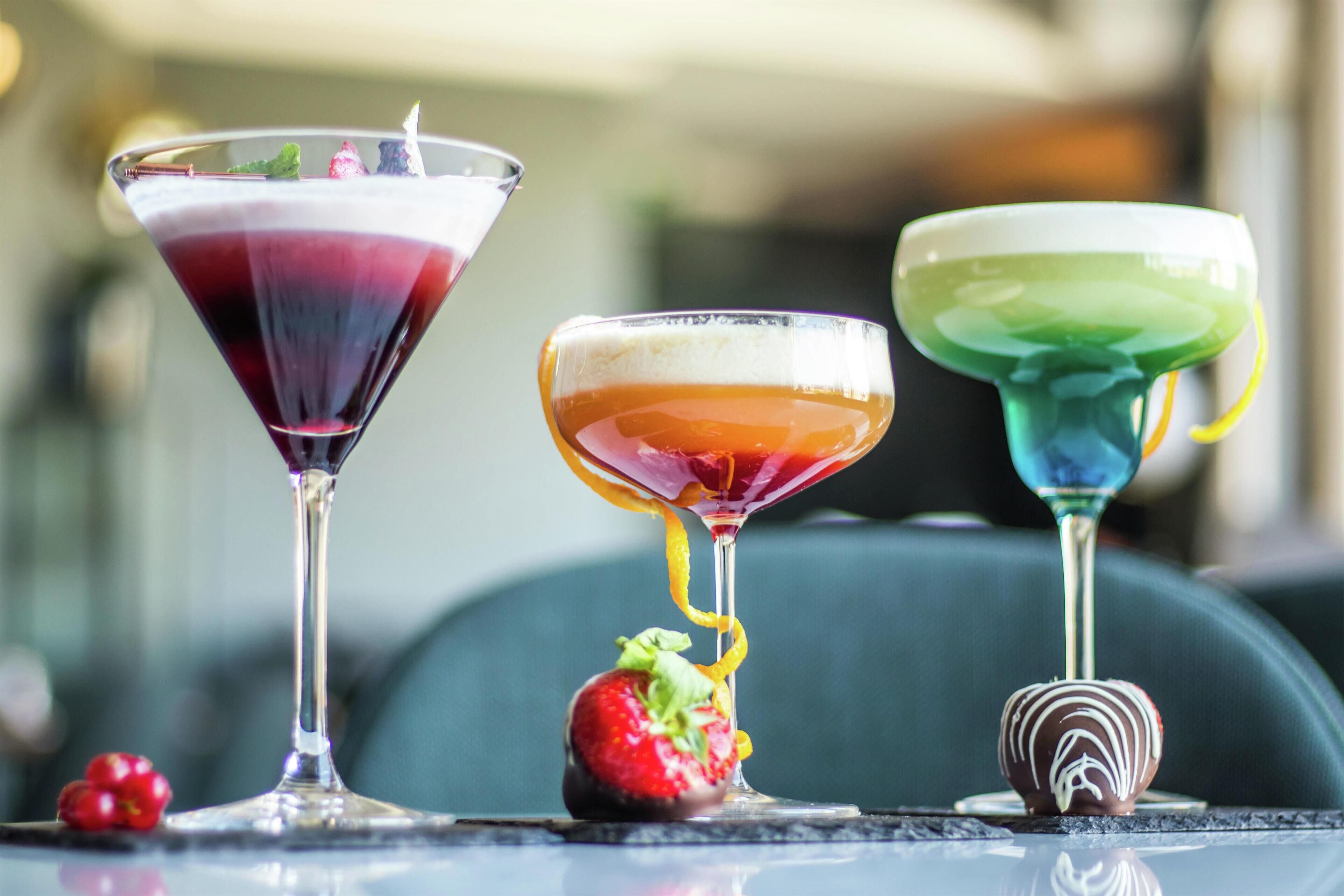 Tre farverige cocktails på et bord med et jordbær og en chokoladedessert.