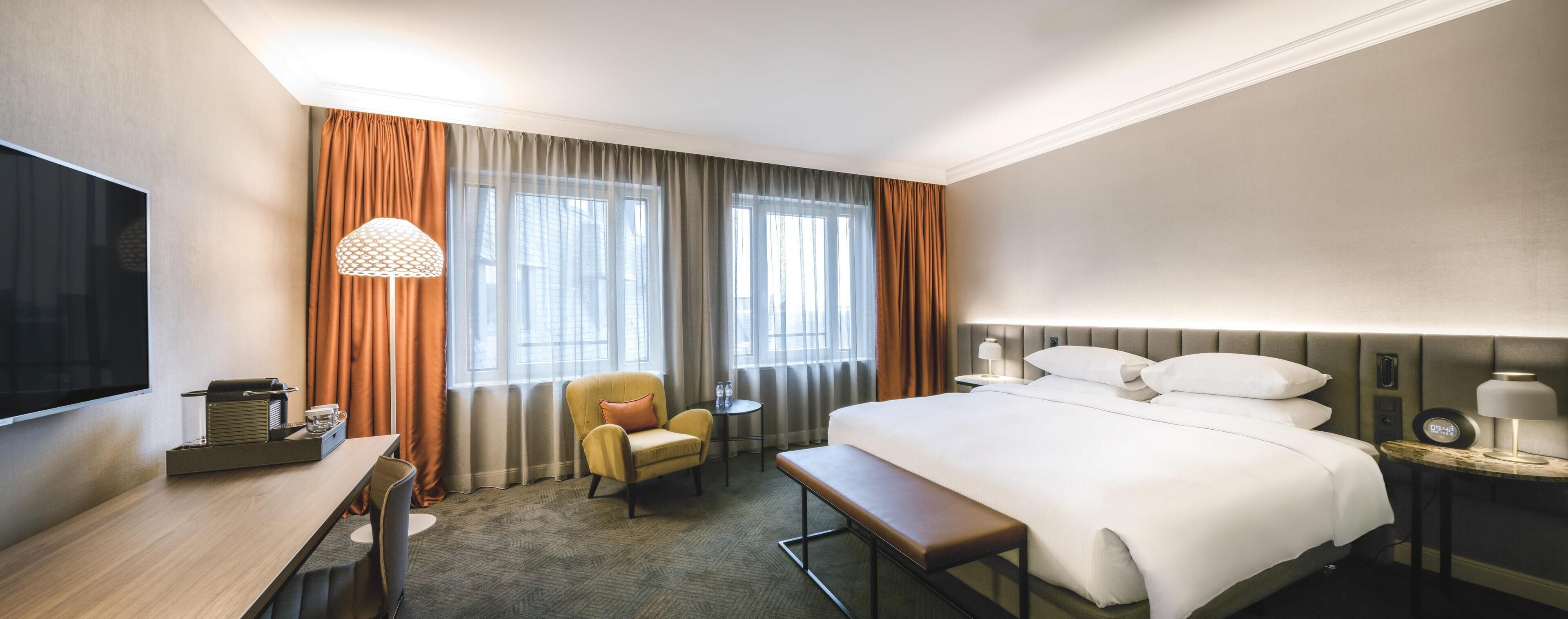 A hotel room with a bed, bedside table, lamp, chair, desk, TV, and curtains.