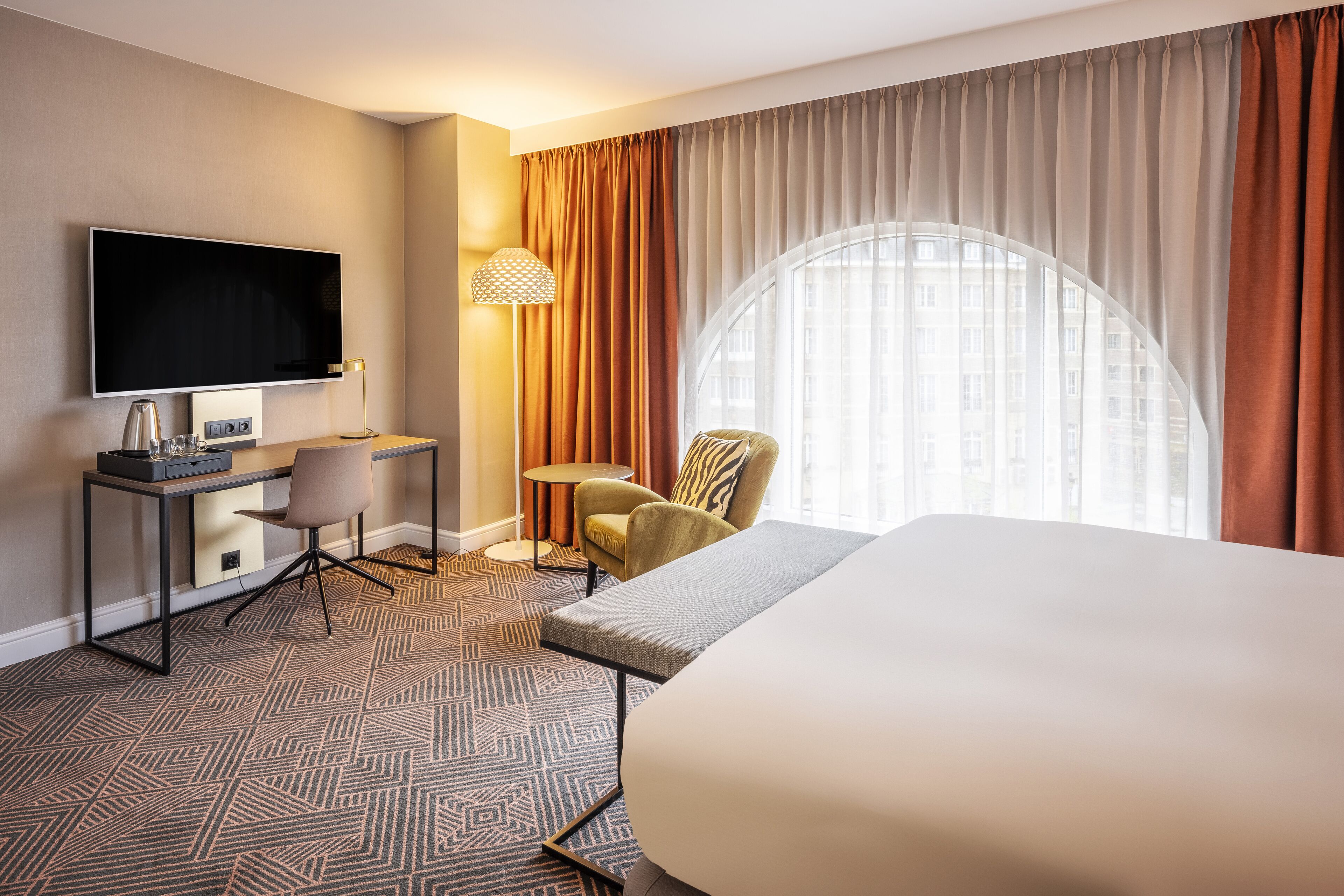 A hotel room with a large bed, a desk with a flat-screen TV, a chair, and a lamp.