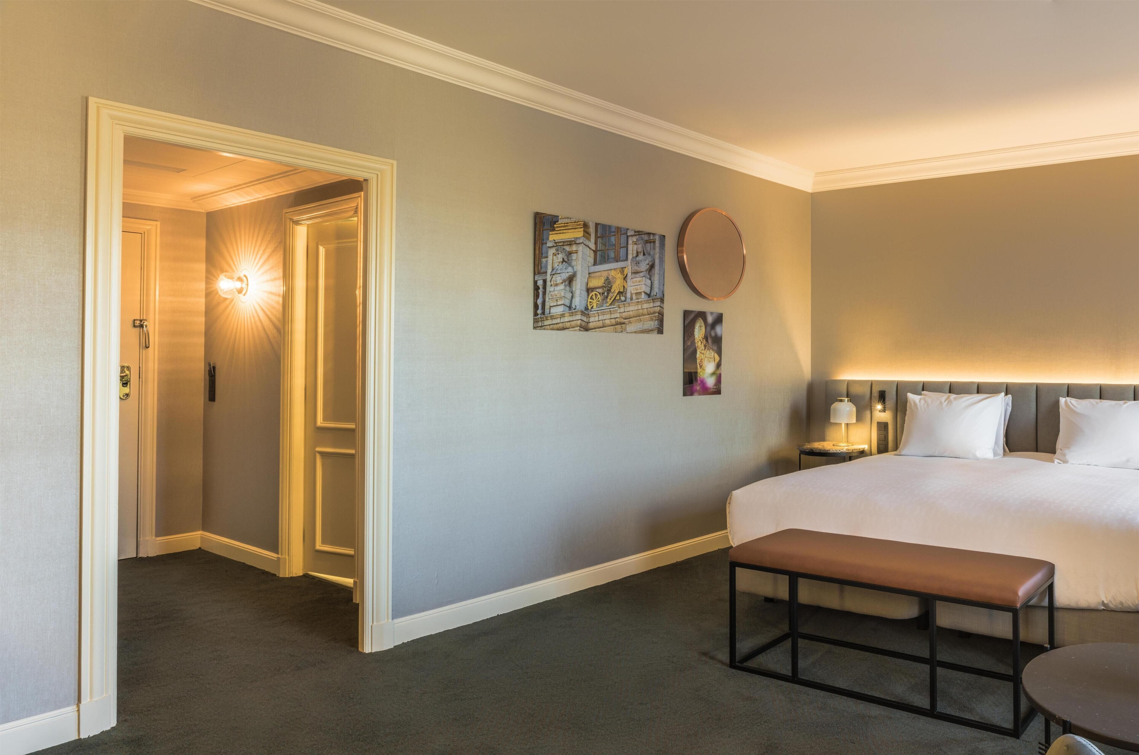 Habitación de hotel con una cama grande, un banco, una mesa redonda, una mesita de noche con lámpara, y una pared adornada con cuadros.