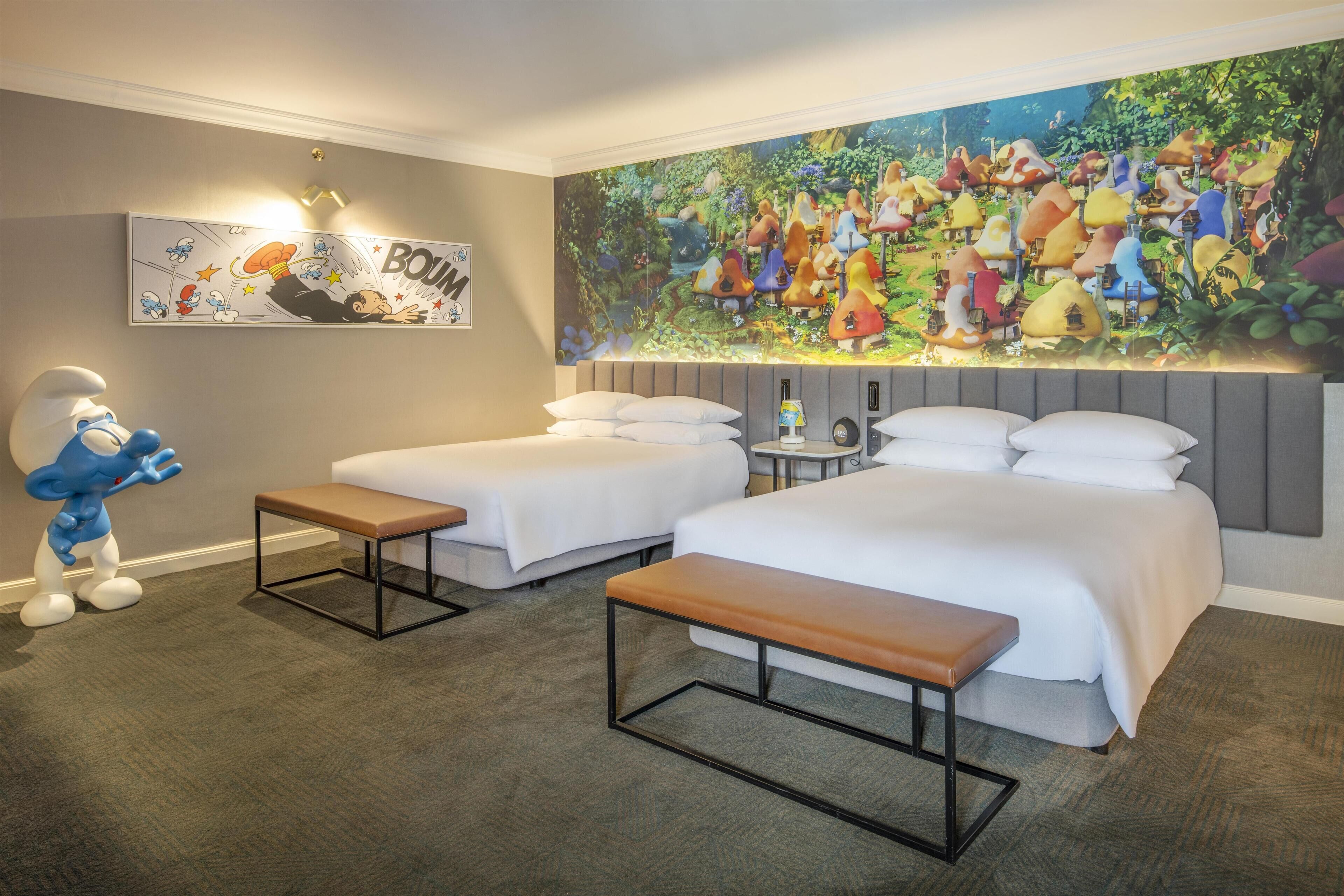 A hotel room with two beds, a bench, a lamp, a small table, and a large mural on the wall.