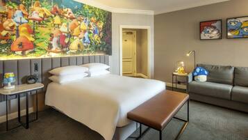 A hotel room with a large mural, a bed, a bench, a sofa, and a small table with a lamp.