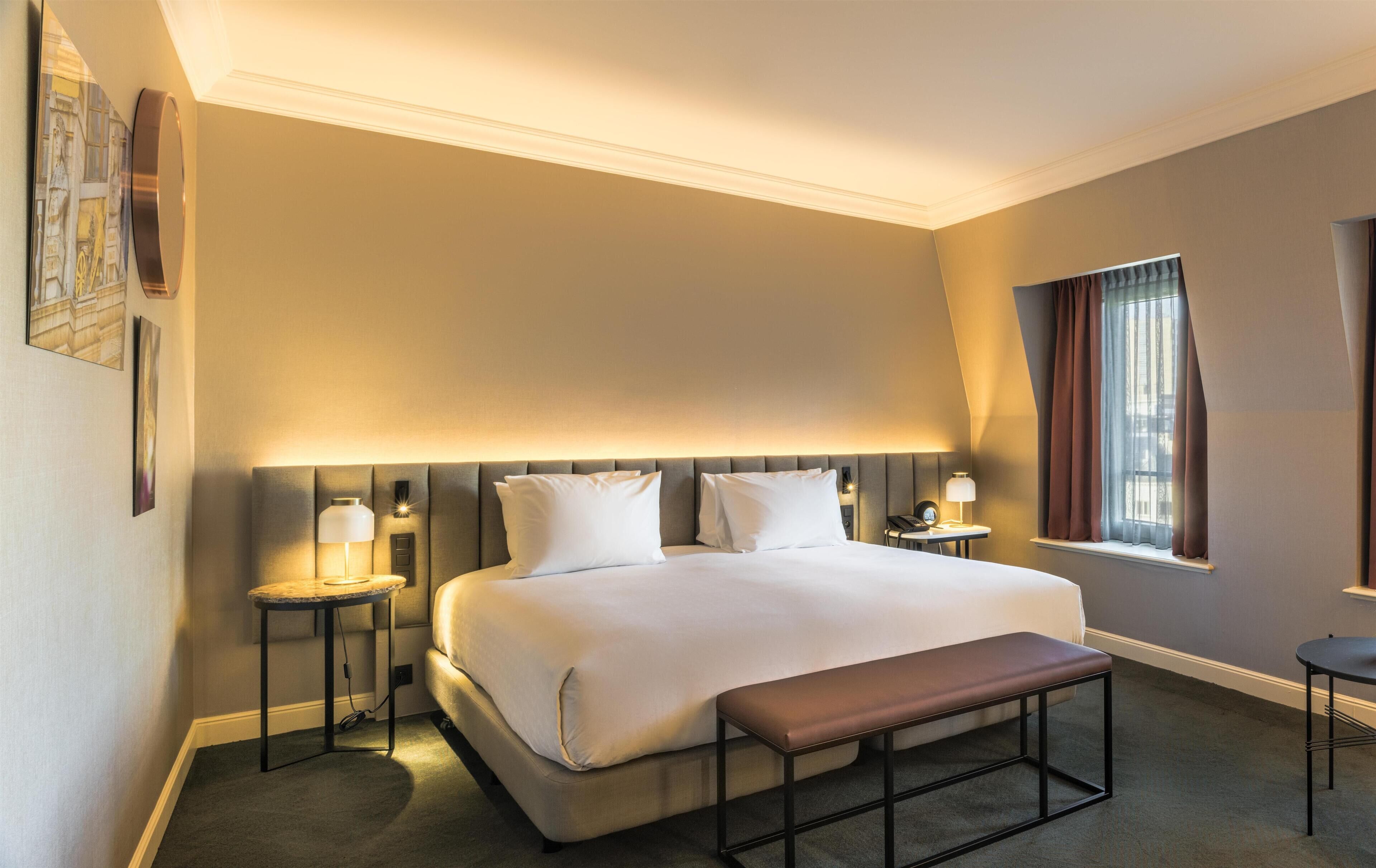 Une chambre d’hôtel avec un grand lit, des tables de chevet, un banc, une fenêtre avec des rideaux et des œuvres d’art accrochées aux murs.
