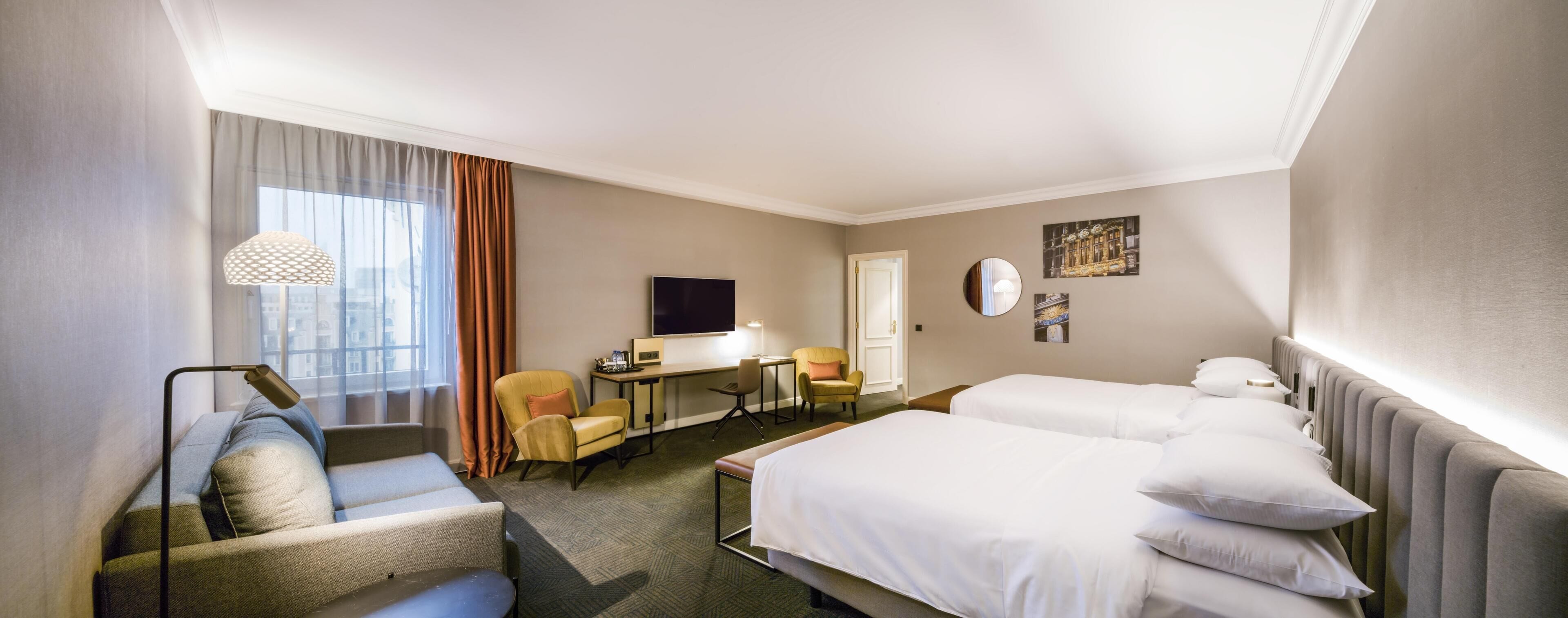 A hotel room with two beds, a desk with a computer, a chair, and a sofa.