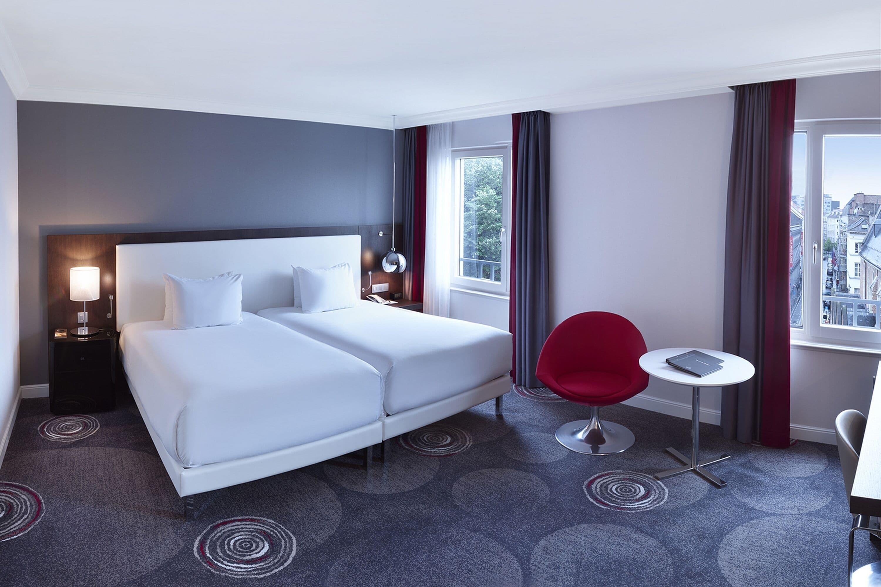 Una habitación de hotel moderna con una cama grande, una silla roja, una mesita y vistas a la ciudad desde la ventana.