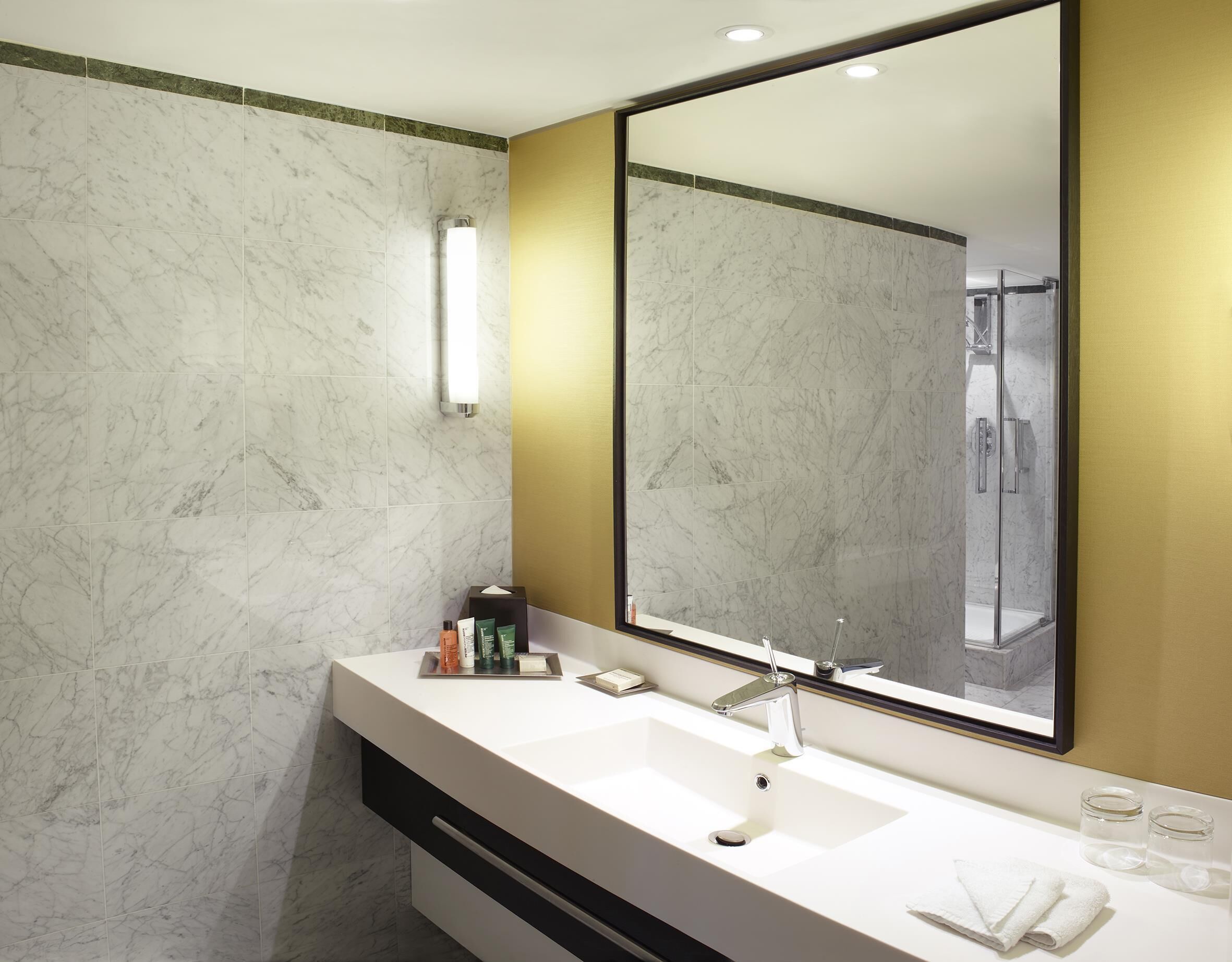 A modern bathroom with a marble wall, a large mirror, a white sink, and a shower area.