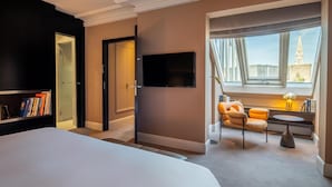 Habitación de hotel con una cama grande, televisor montado en la pared, zona de estar con una silla y una mesa, y ventana con vistas a un edificio.
