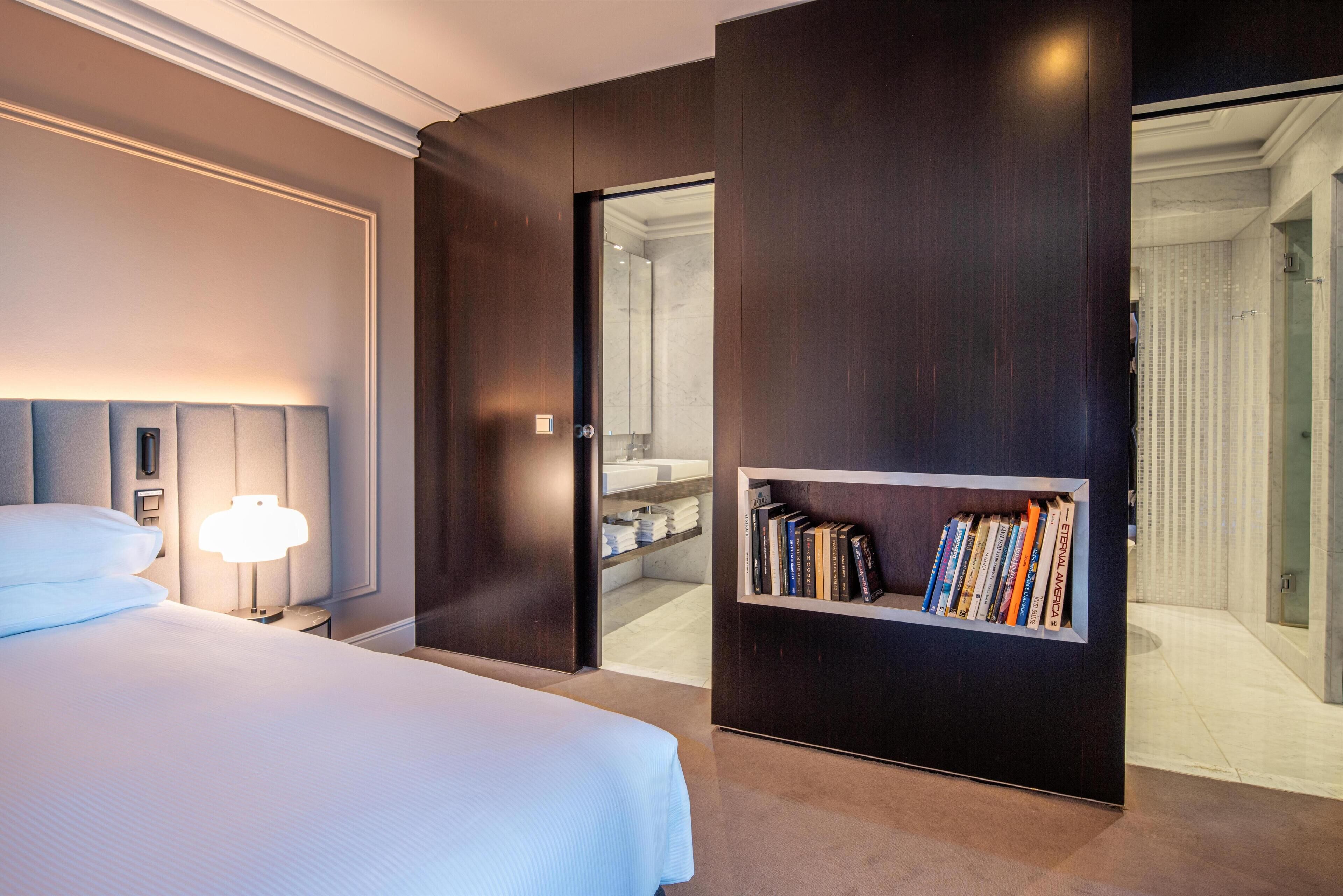 A modern hotel room with a neatly made bed, a bedside lamp, and a bookshelf with books.