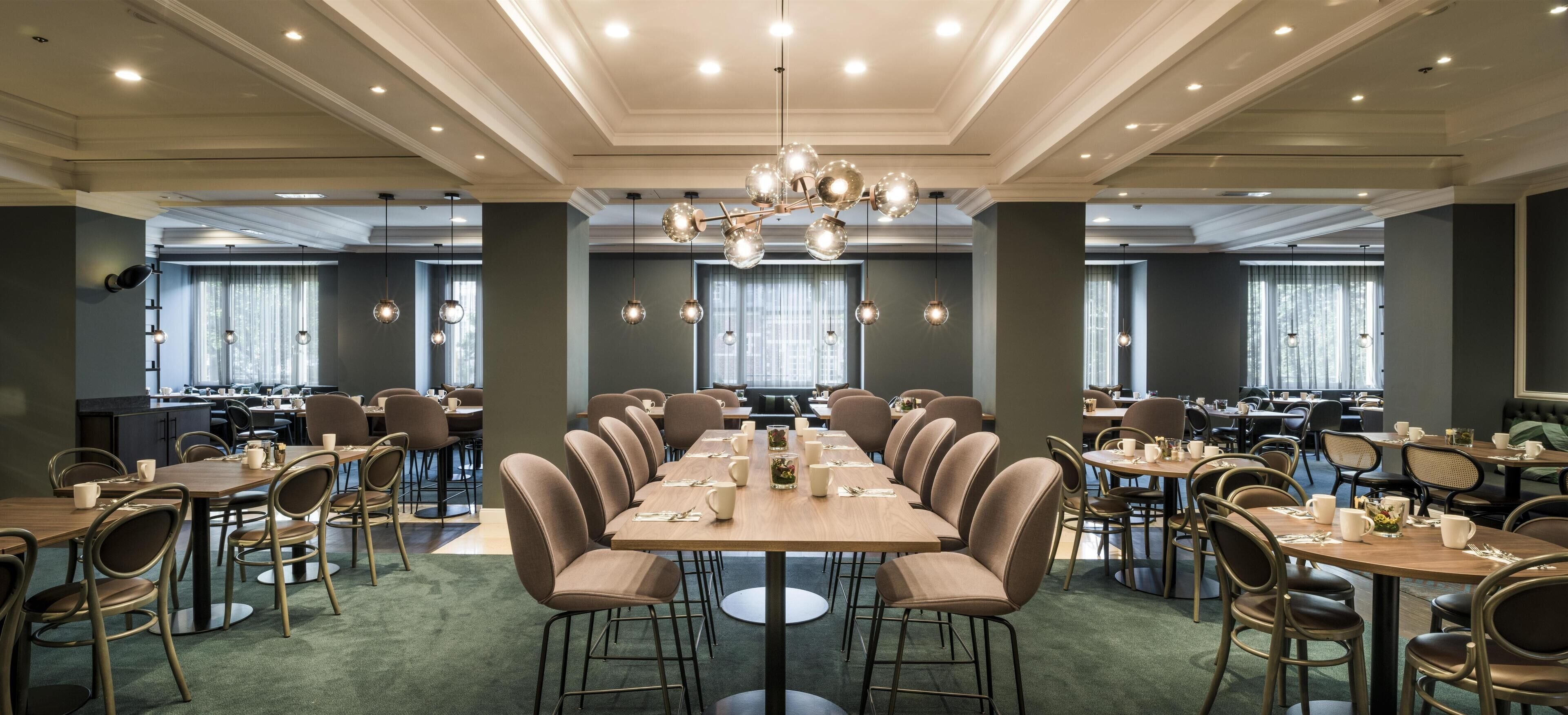 Un amplio comedor con numerosas mesas y sillas, un candelabro y ventanas grandes con cortinas.