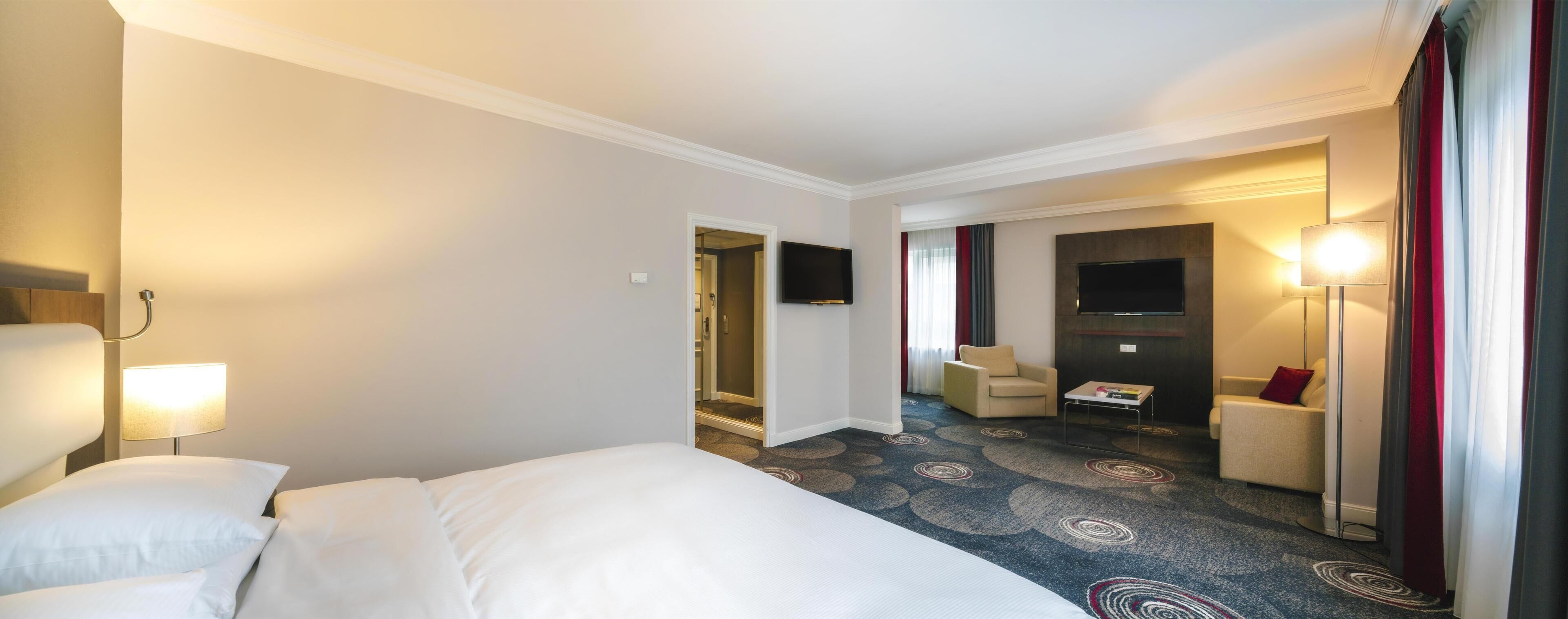 A hotel room with a bed, a desk, a chair, a TV, and a carpeted floor.