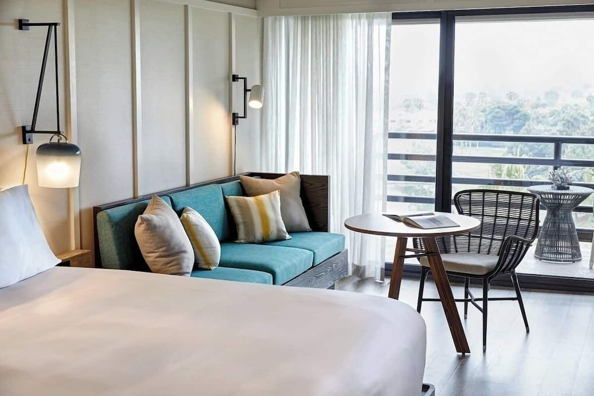 A hotel room with a bed, a sofa, a small round table, two chairs, a lamp, and a large window with a view.