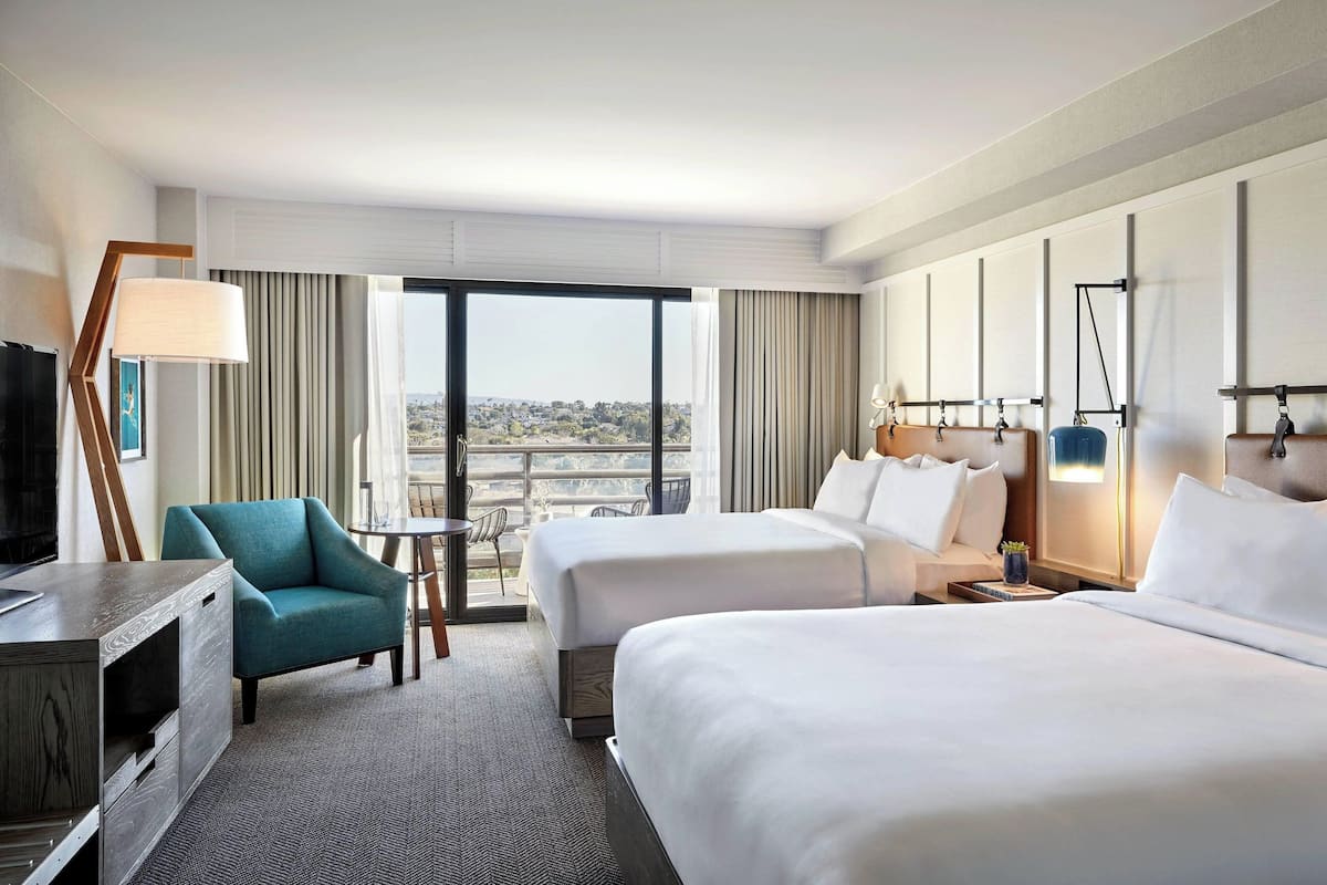 A hotel room with two beds, a desk, a chair, a TV, and a large window offering a city view.