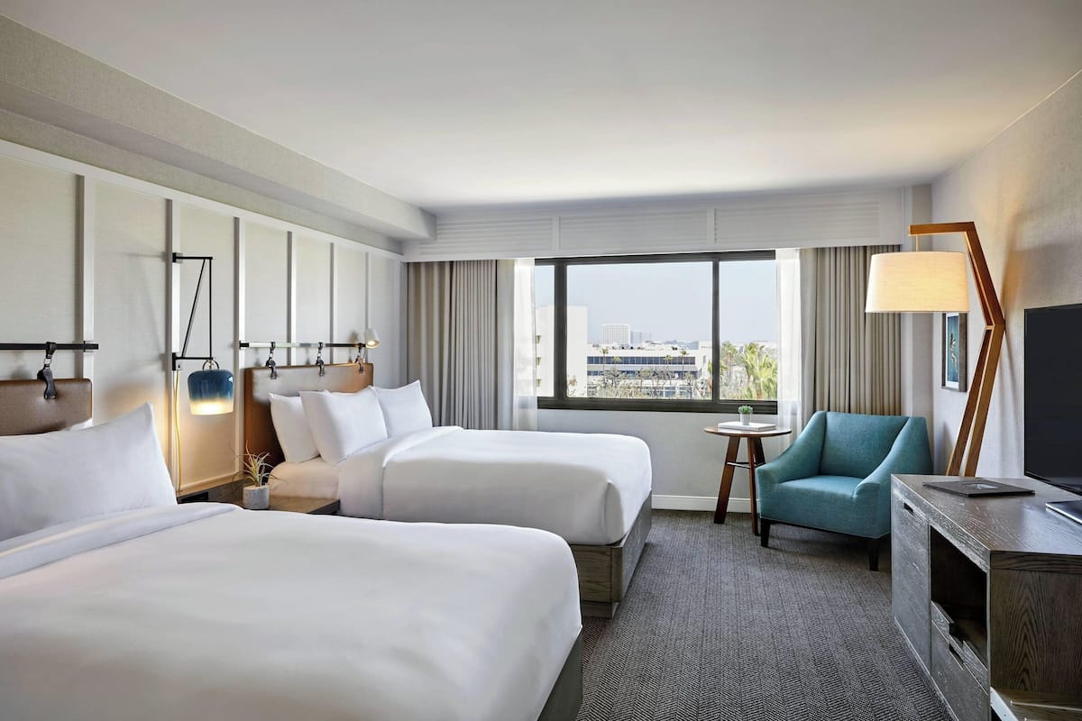 A hotel room with two beds, a TV, a desk, and a view of the city.