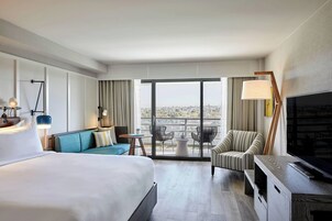 Habitación de hotel con una cama grande, un sofá, un televisor, un escritorio y un balcón con vistas a la ciudad.