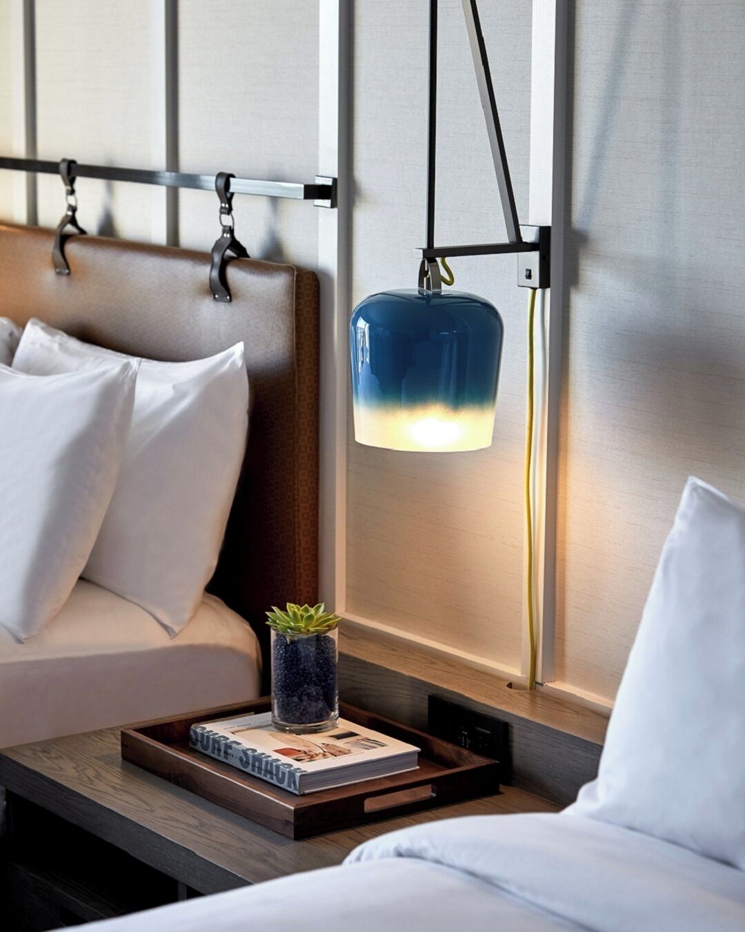 Moderni hotellihuone, jossa on sohva, yöpöytä, jossa on kirja ja kasvi, sekä riippuvalaisin.