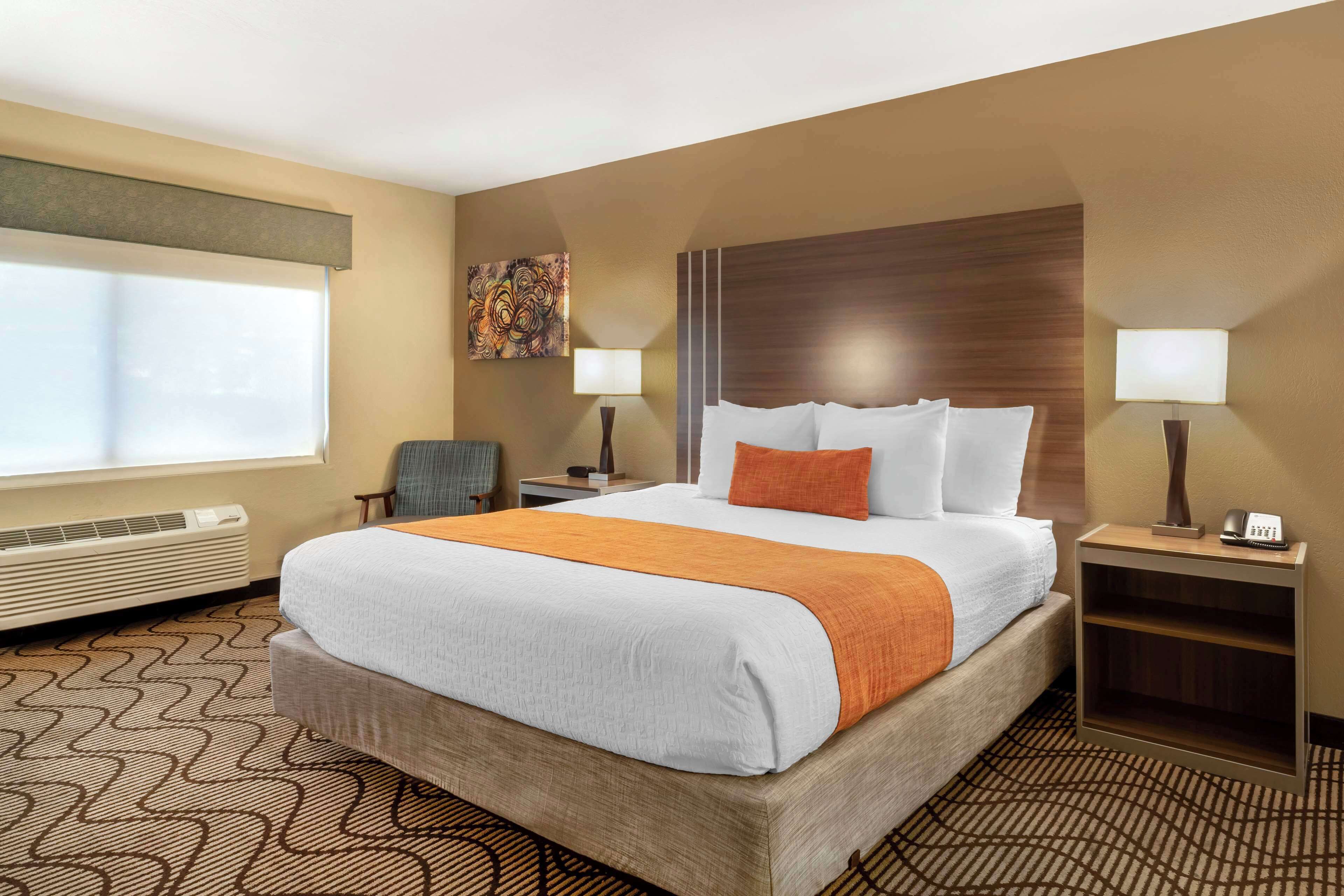 A hotel room with a large bed, two bedside tables, a chair, a window with blinds, a wall-mounted artwork, and a patterned carpet.