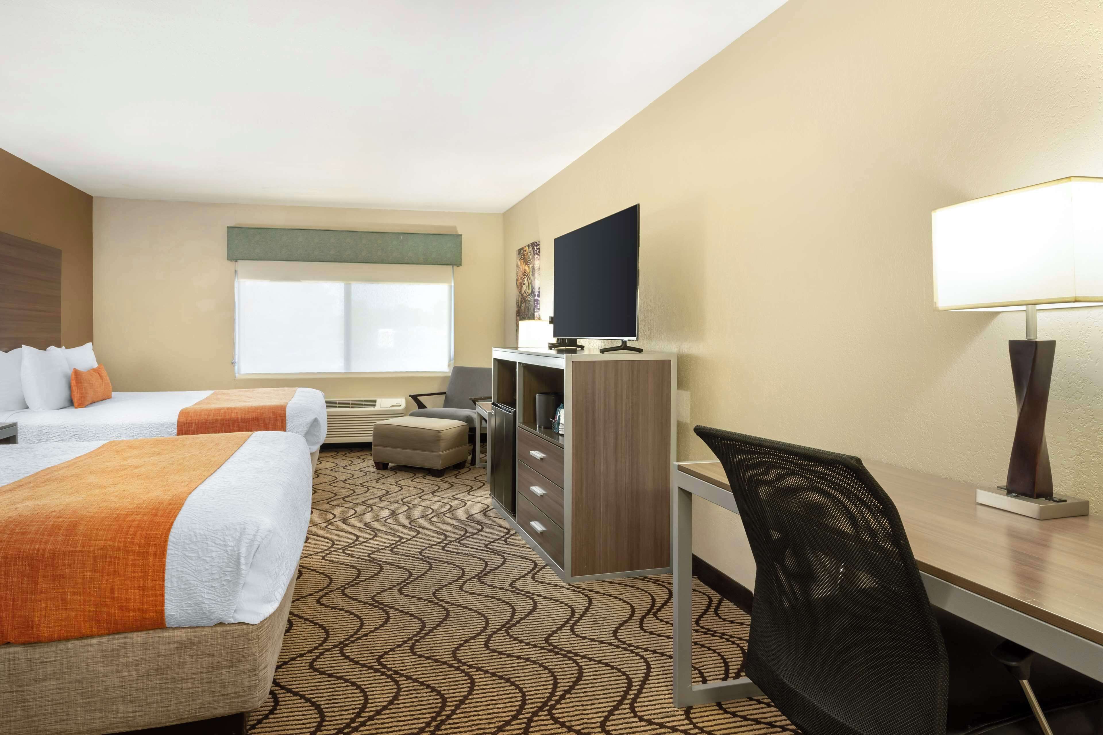 A hotel room with two beds, a desk, a chair, a TV, and a lamp.