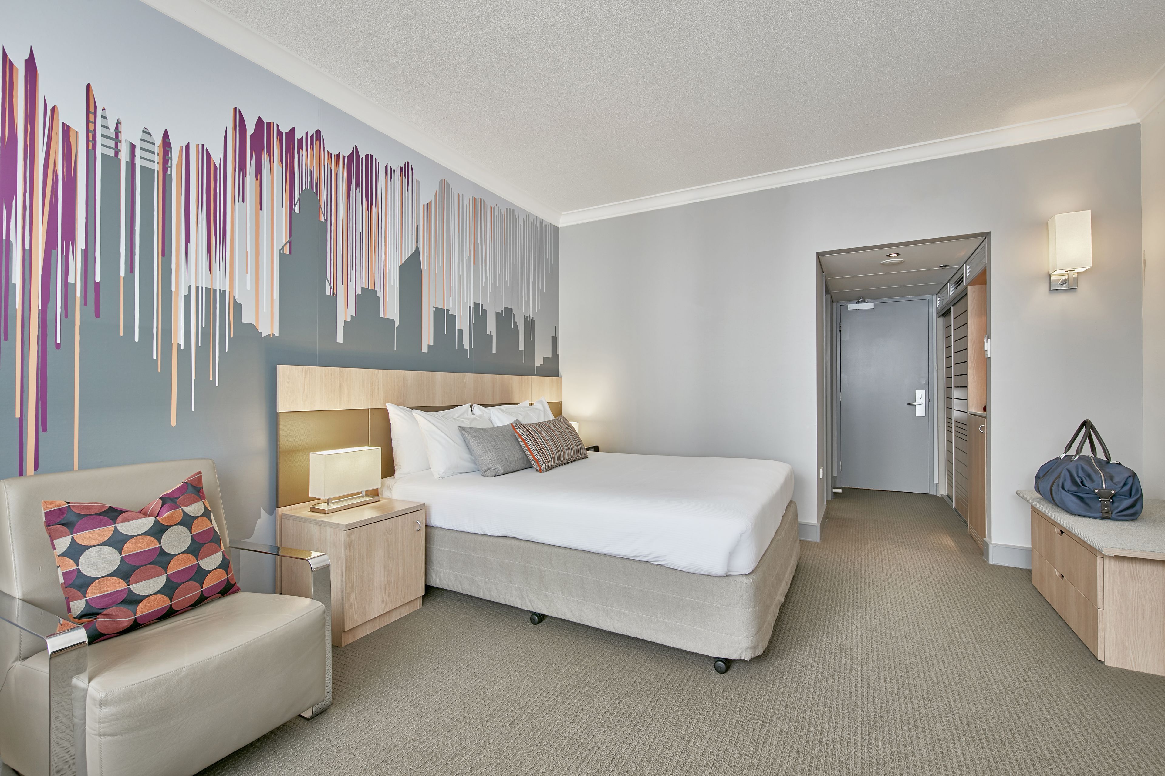 A hotel room with a bed, a chair, a nightstand, a wall art piece, and a hallway leading to a closet.