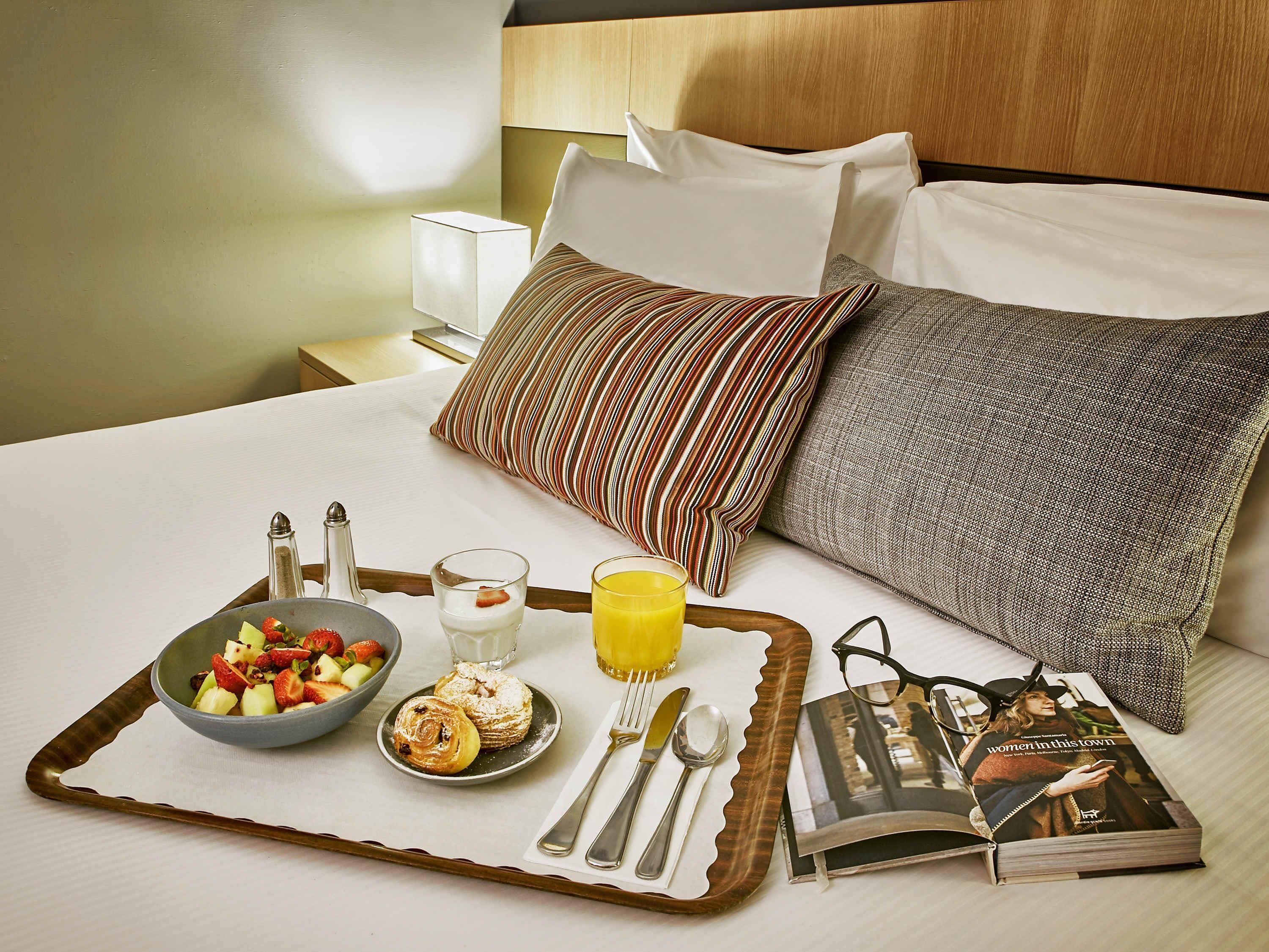 A neatly made bed with pillows, a tray with breakfast including fruit, pastries, and juice, and a book on the side.