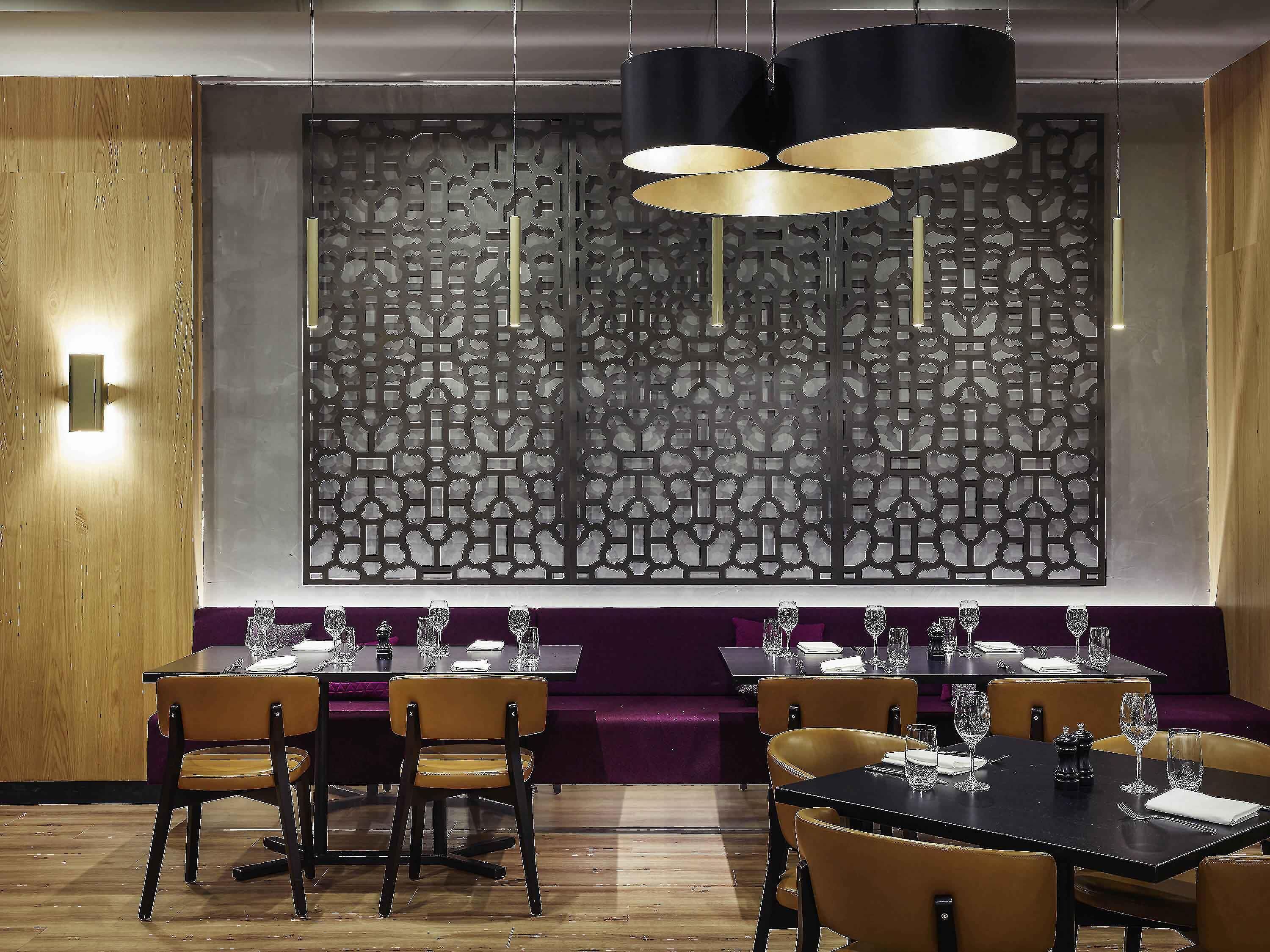 A restaurant interior with dark wood paneling, a patterned wall, and black and gold pendant lighting. Tables are set with wine glasses and cutlery.