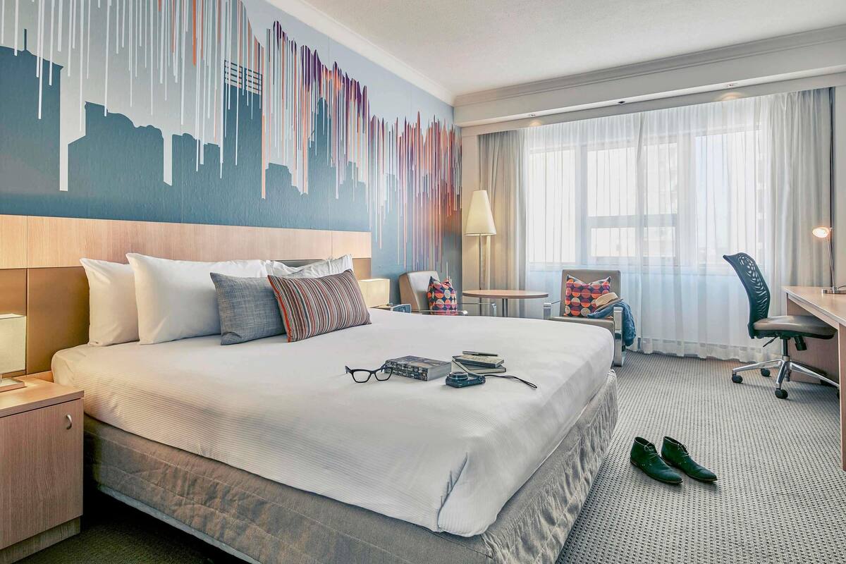 Ein Hotelzimmer mit einem großen Bett, einem Schreibtisch, einem Sessel und einem Wandgemälde mit Stadtansicht.