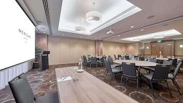Un salón de conferencias con una gran mesa, sillas, una pantalla de proyector y un orador.