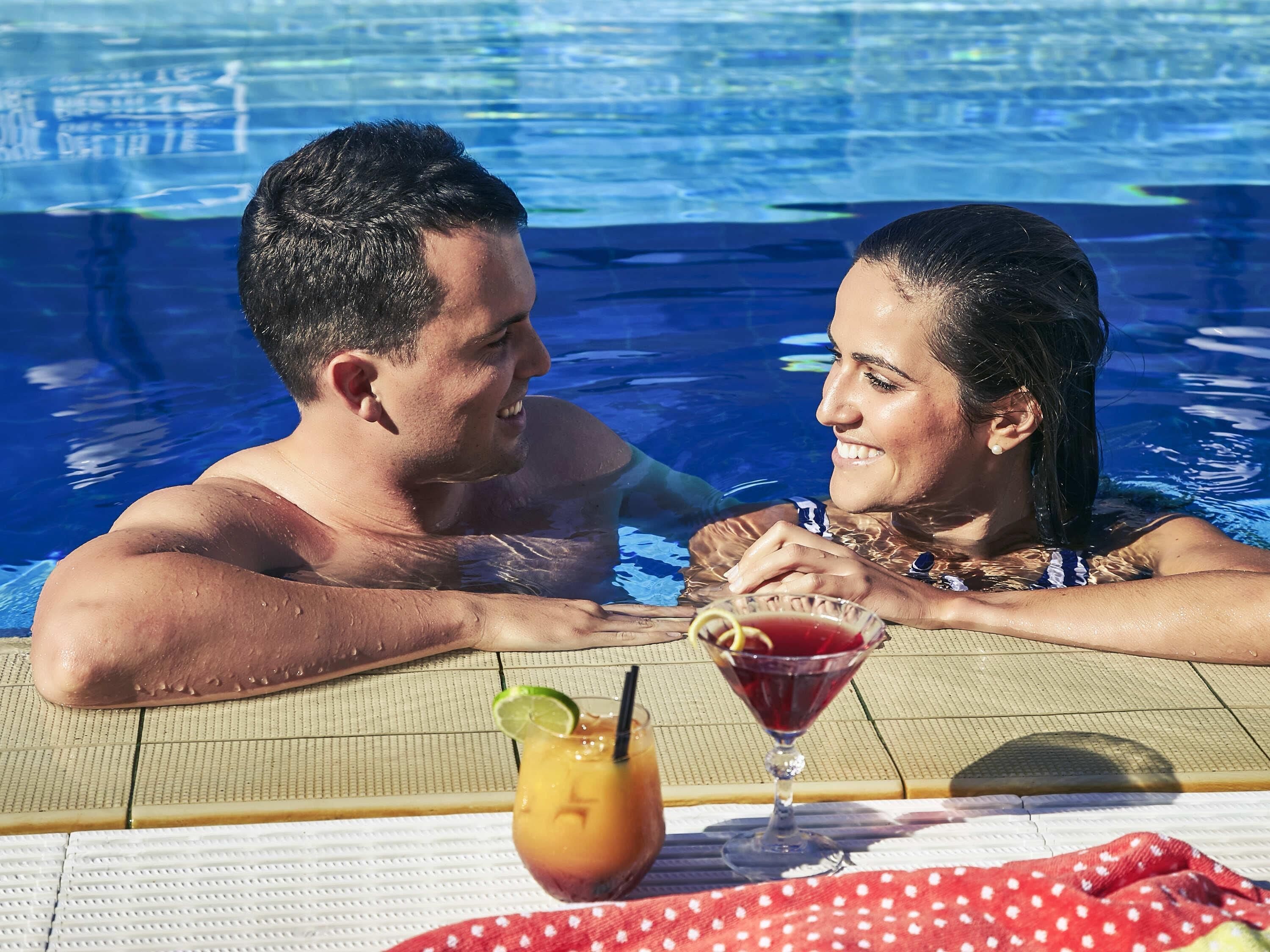 A couple in a swimming pool with drinks on a table.