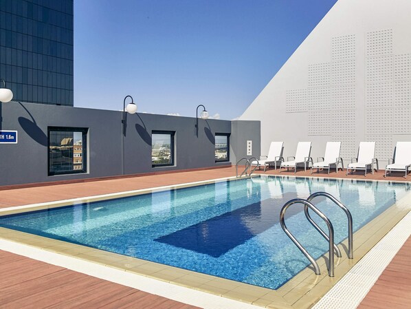 A modern indoor swimming pool with clear blue water, surrounded by lounge chairs and a view of a cityscape through windows. - Mercure Perth (Perth)