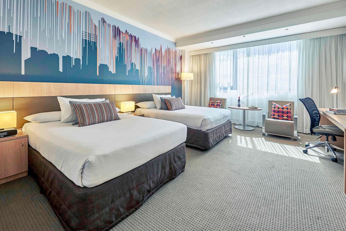 A hotel room with two beds, a desk, a chair, a small table, and a cityscape mural on the wall.