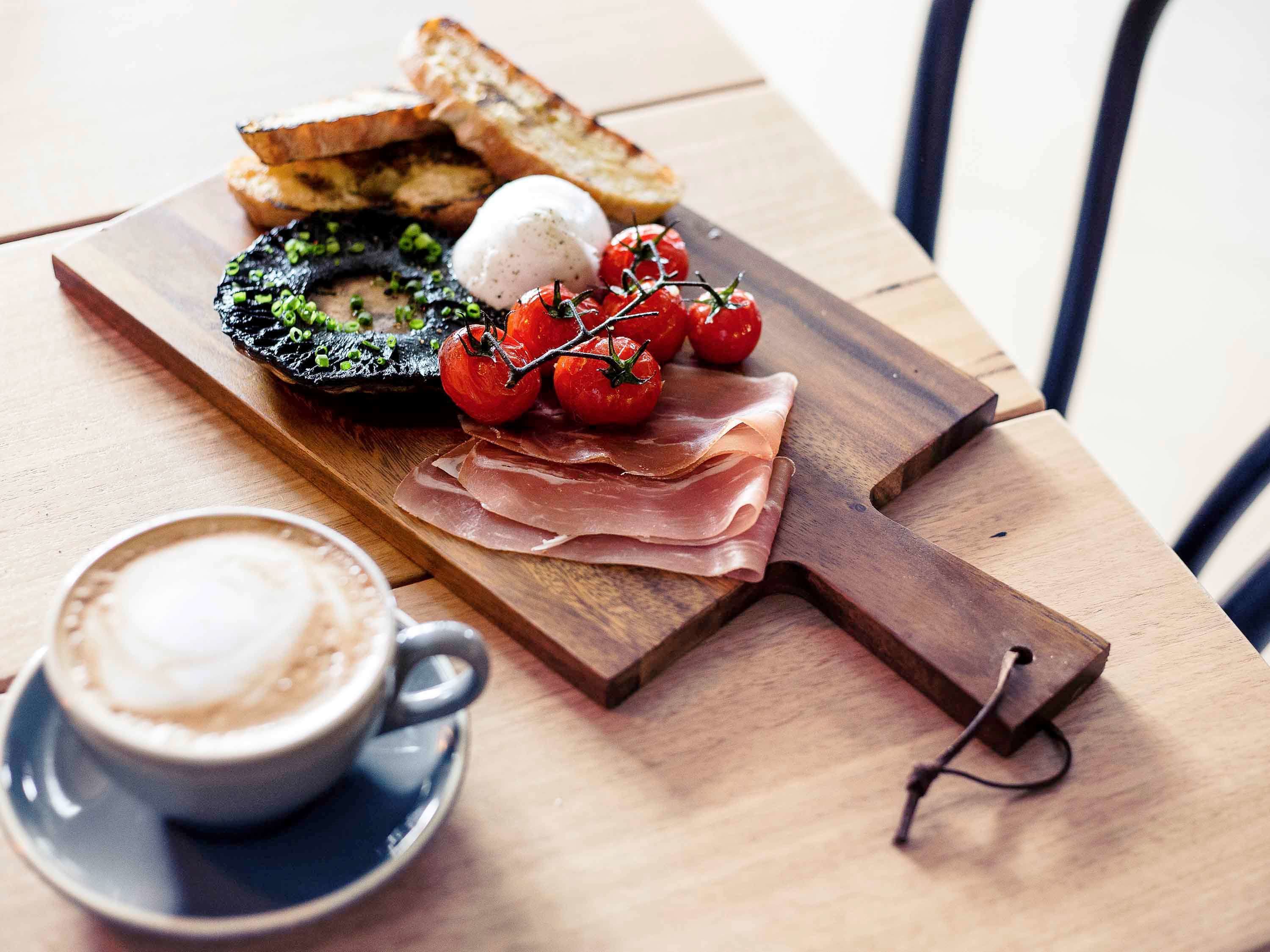 A wooden board with prosciutto, cherry tomatoes, a poached egg, and toasted bread on a table.