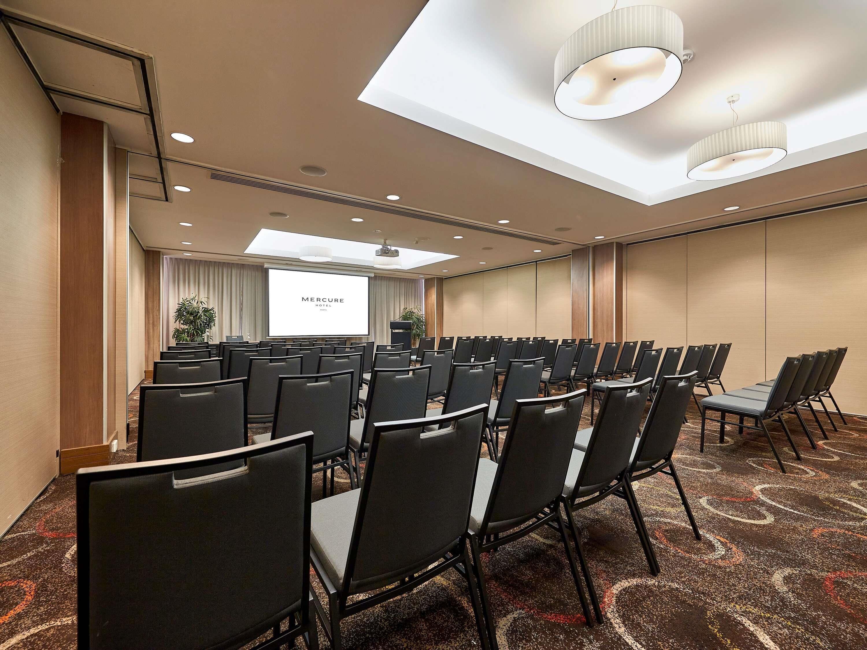 A conference room with rows of chairs, a projector screen, and a ceiling light fixture.