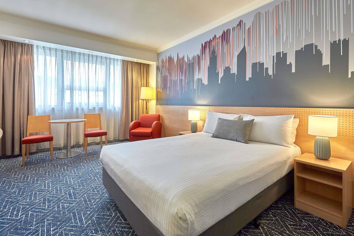 Ein Hotelzimmer mit einem großen Bett, zwei Sesseln, einem kleinen Tisch, einem Nachttisch und einem Wandgemälde mit Stadtansicht.