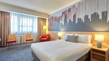 A hotel room with a large bed, two chairs, a small table, a nightstand, and a cityscape mural on the wall.