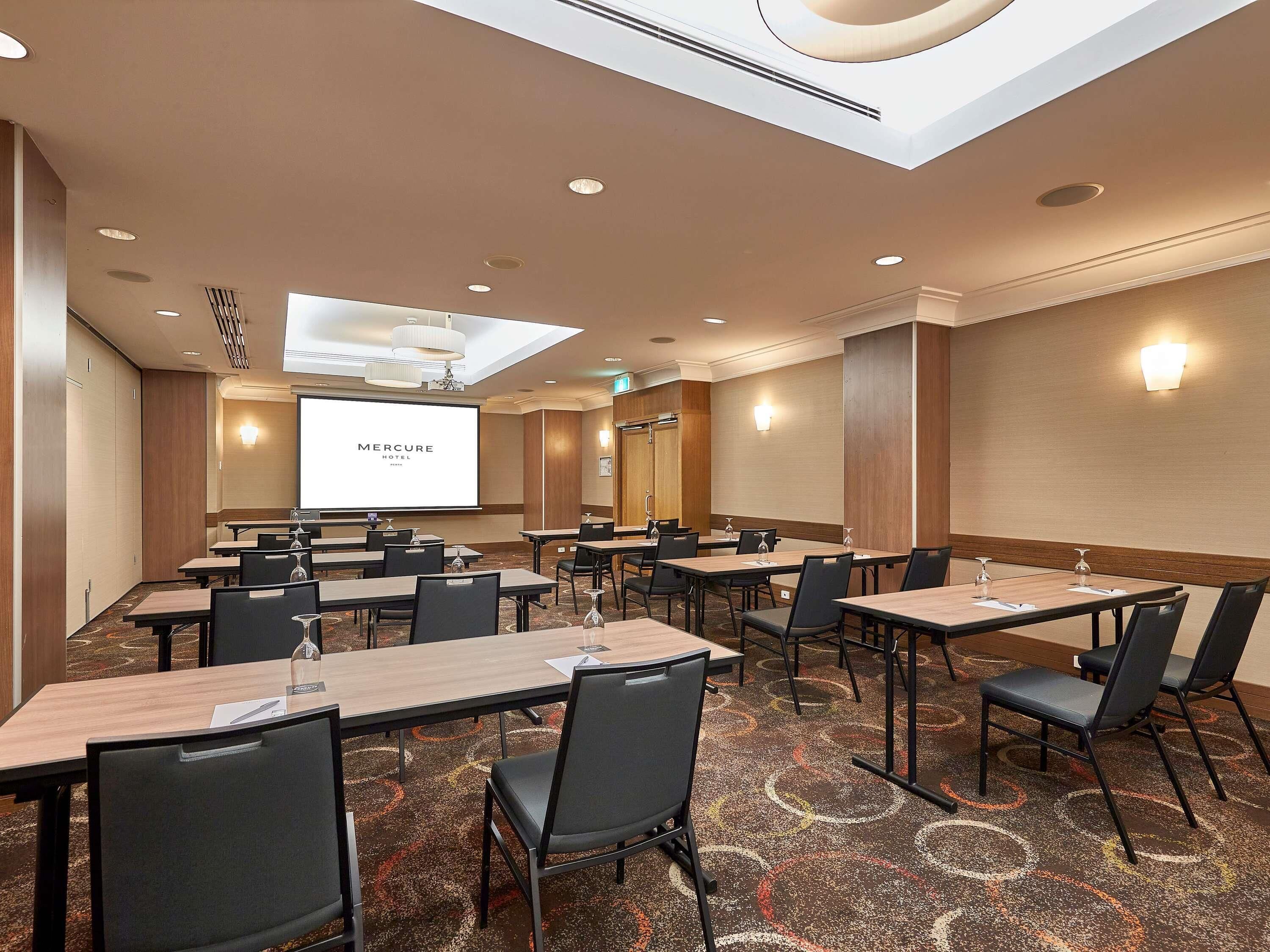 Una sala conferenze con diversi tavoli e sedie, uno schermo che mostra 'Mercure Hotel' e un soffitto con lucernari.