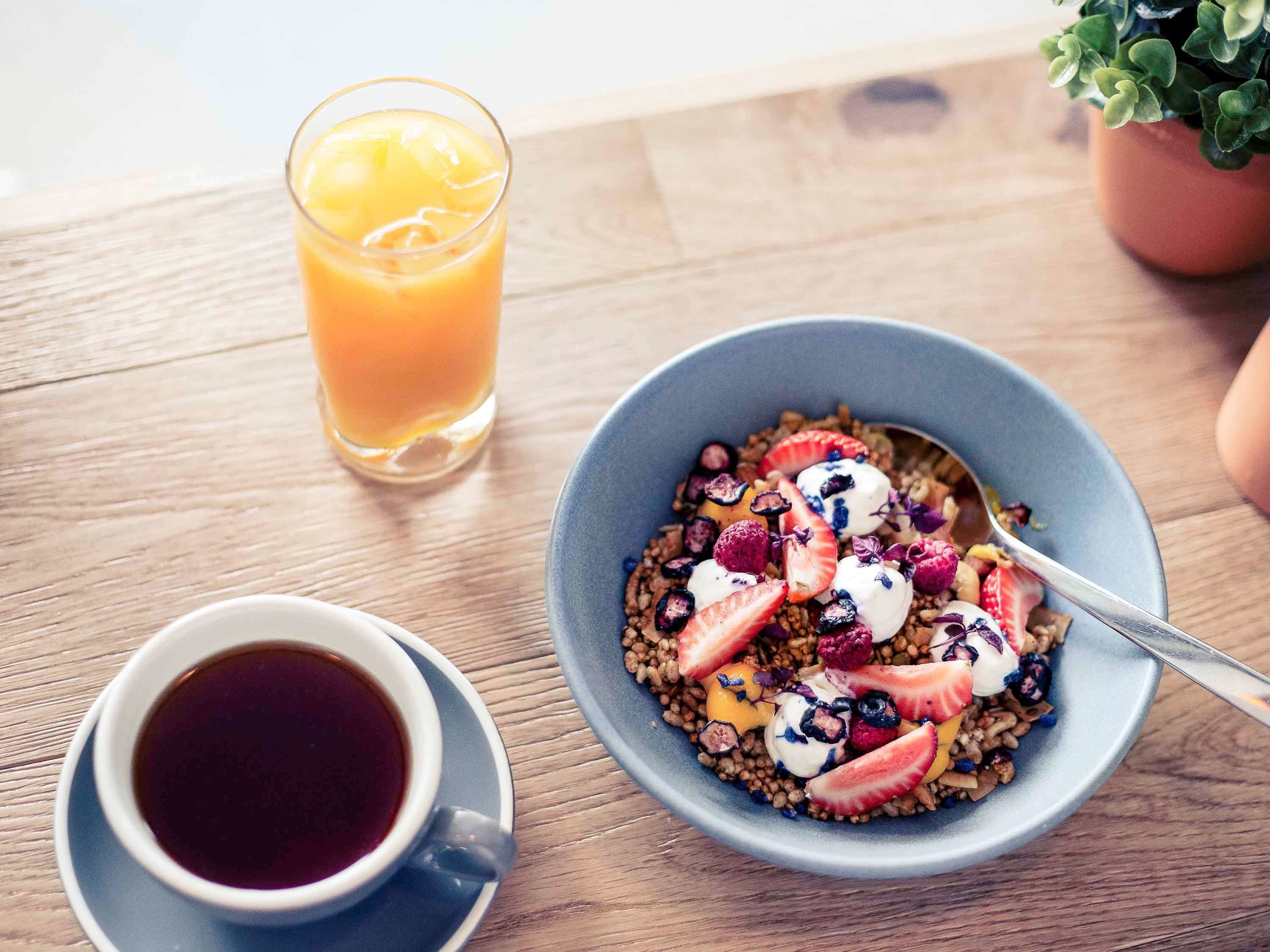 A breakfast setting with a bowl of cereal topped with fruits and yogurt, a glass of orange juice, and a cup of coffee on a wooden table.