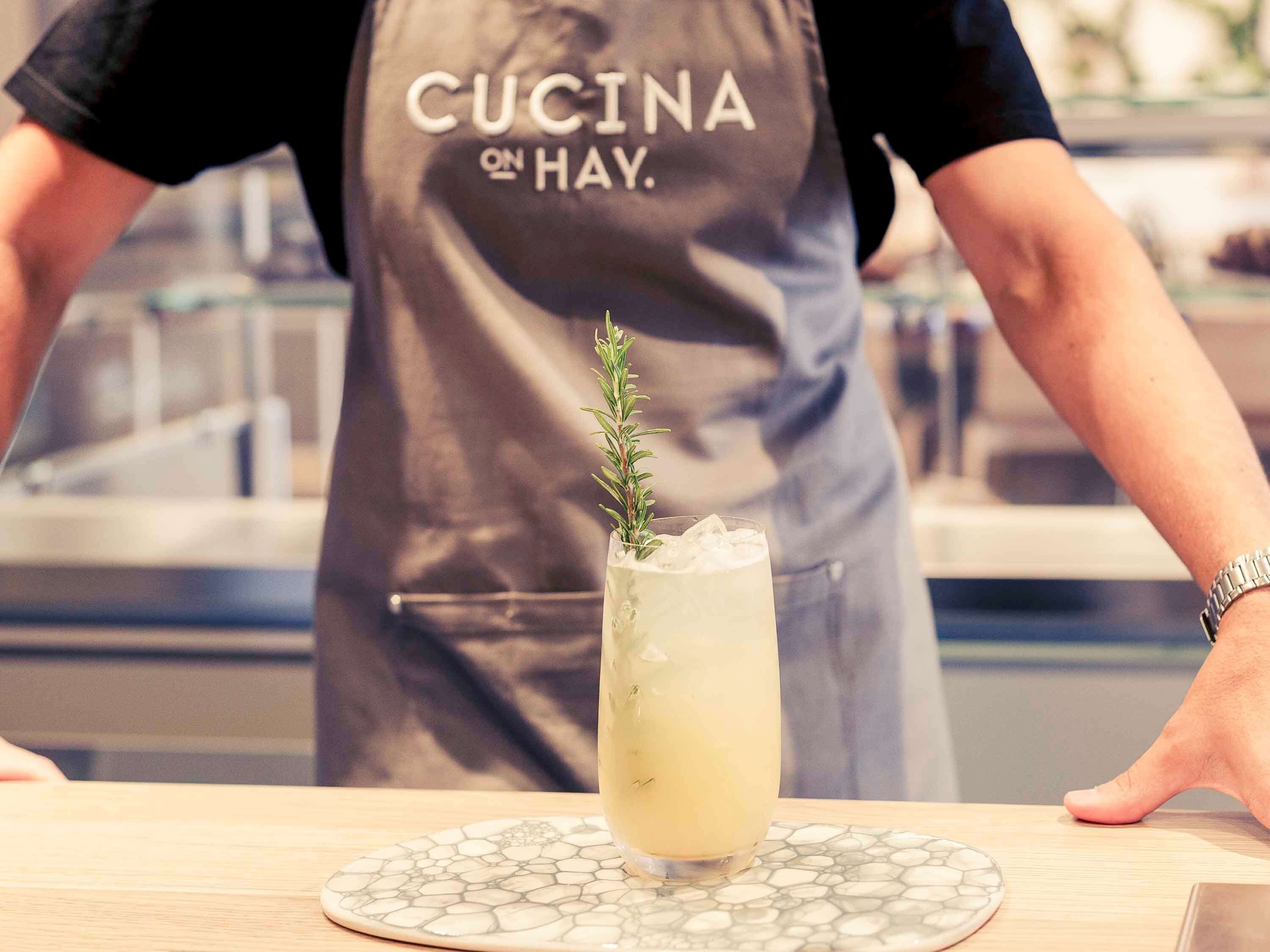 En person i forklæde med teksten "CUCINA ON HAY", der serverer en drik med en rosmarinkvist på en underkop.